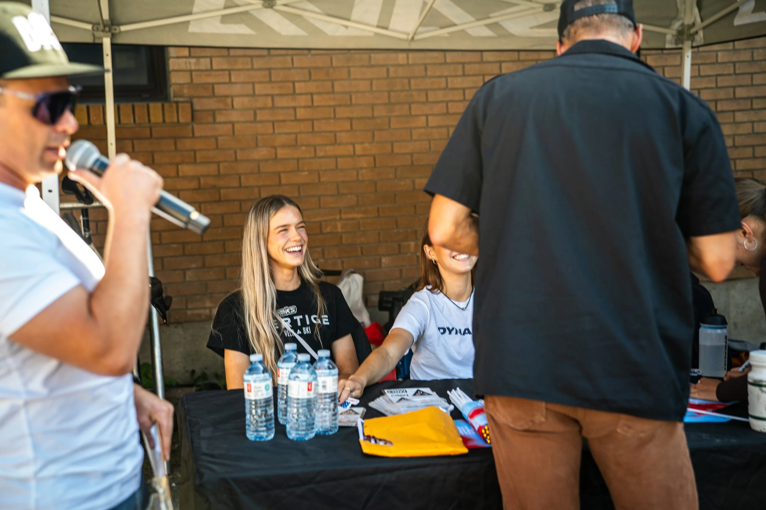 Two women sitting at a table with water bottles and flyers, smiling and interacting with a person standing in front of them during an outdoor event under a canopy.