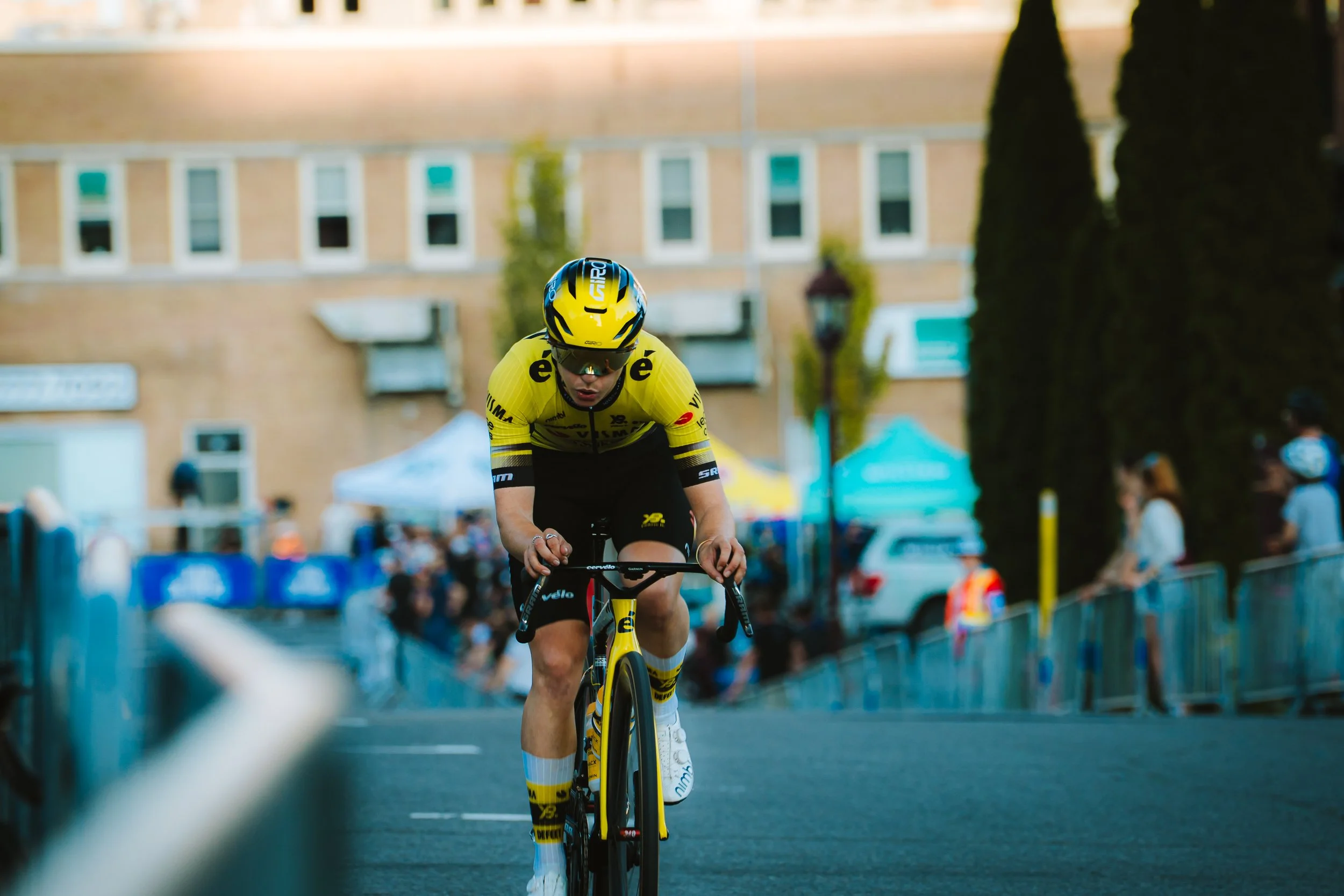 Cyclist in yellow and black uniform riding a bicycle during a race, with spectators and tents in the background.