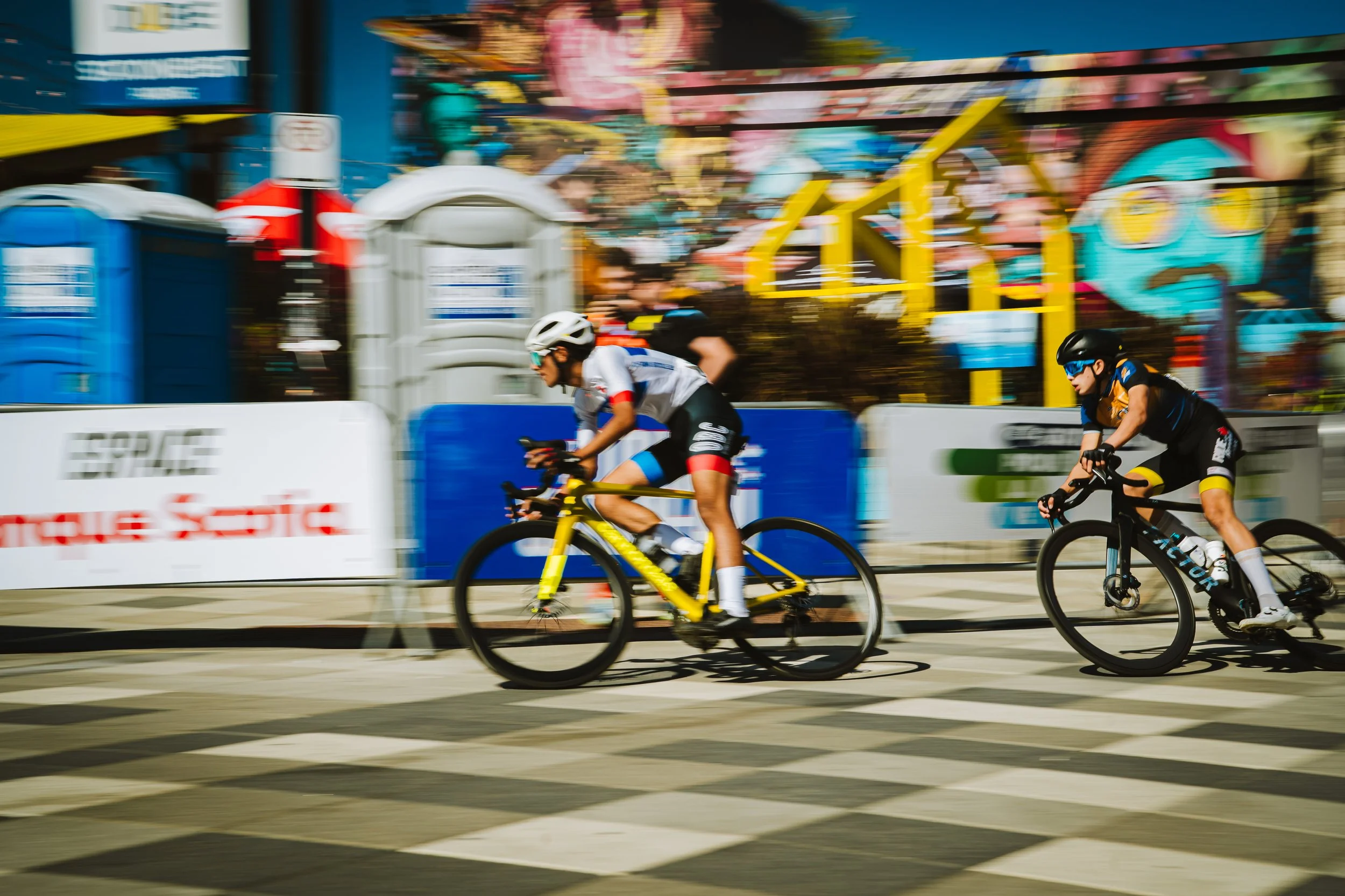 Two cyclists racing on a city street with colorful murals and portable toilets in the background.