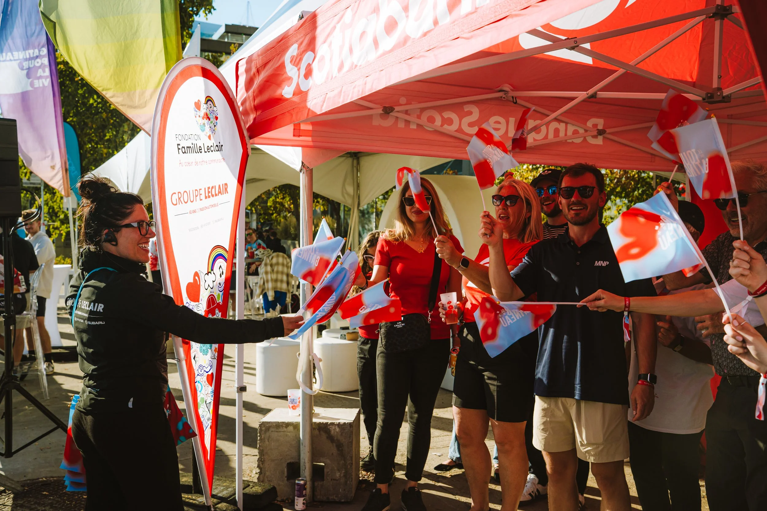 Group of people celebrating under a red canopy at an outdoor event, waving small flags with a smiling woman handing out flags. There are colorful banners and trees in the background.