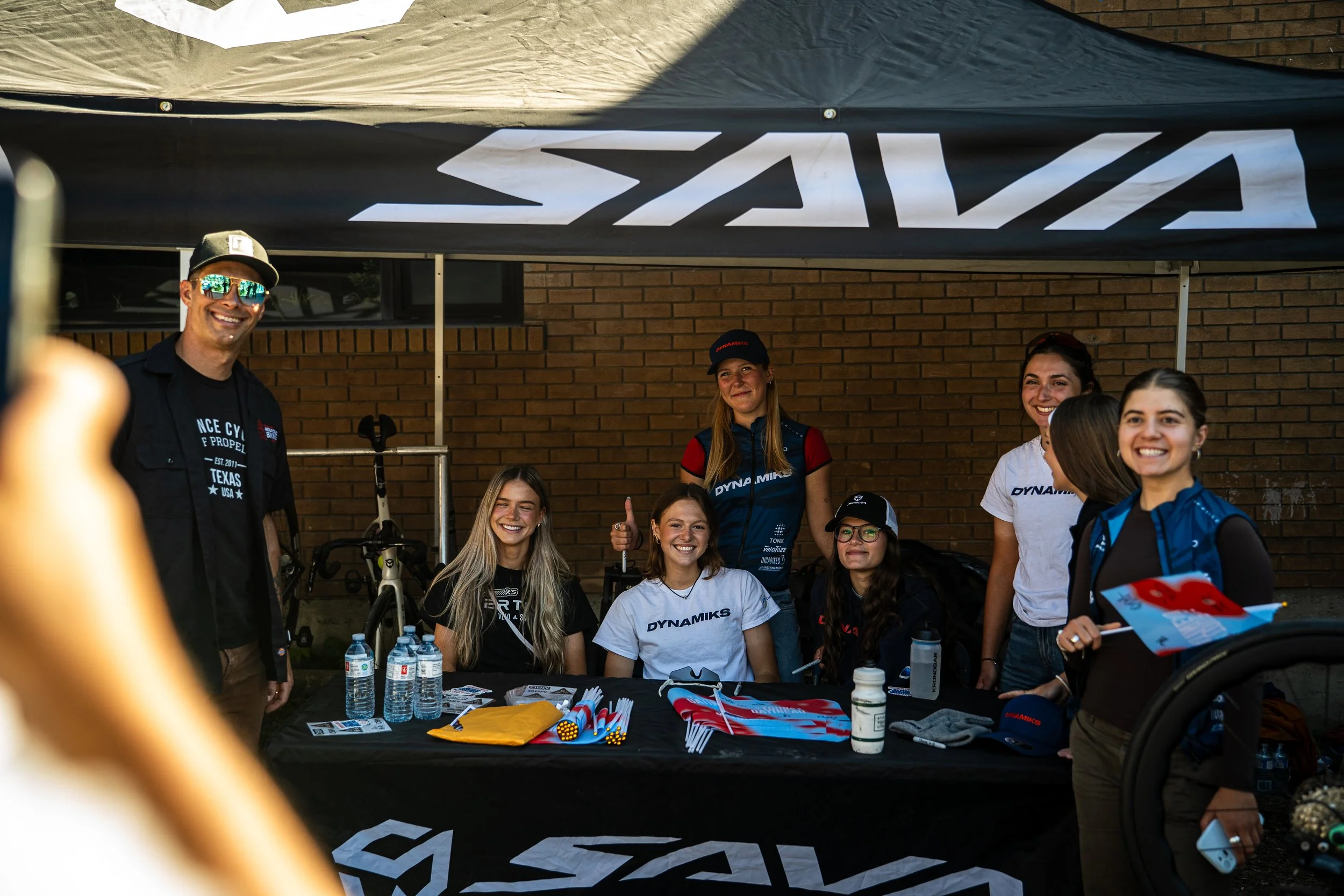 Group of people at a promotional event or booth, sitting and standing behind a table with branded merchandise, water bottles, and flyers, outdoors with a brick building in the background.