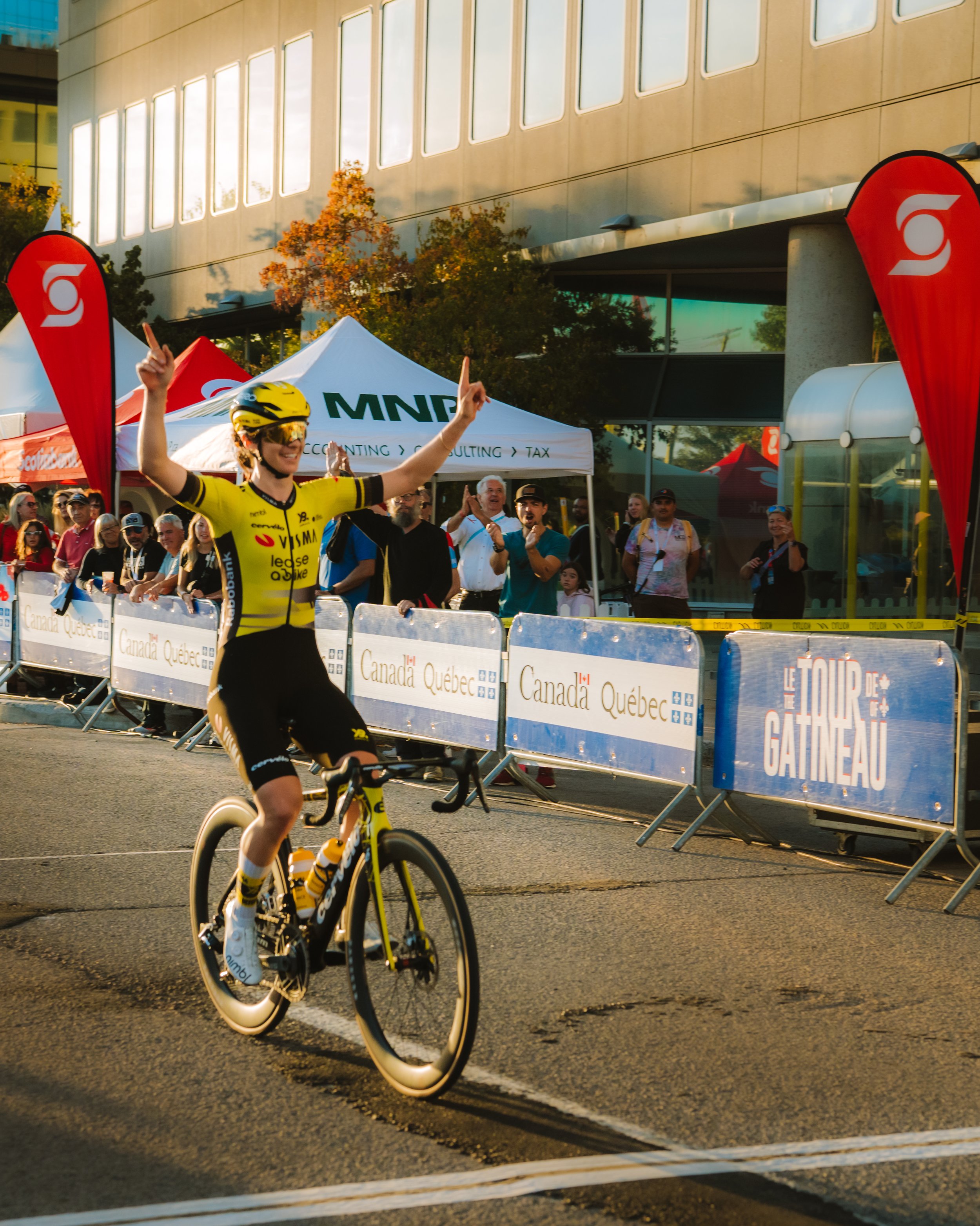 A cyclist in yellow and black riding a bike across the finish line at a cycling event, with people cheering and taking photos behind barriers marked with signs for Canada Quebec and Le Tour des Gatsneau.