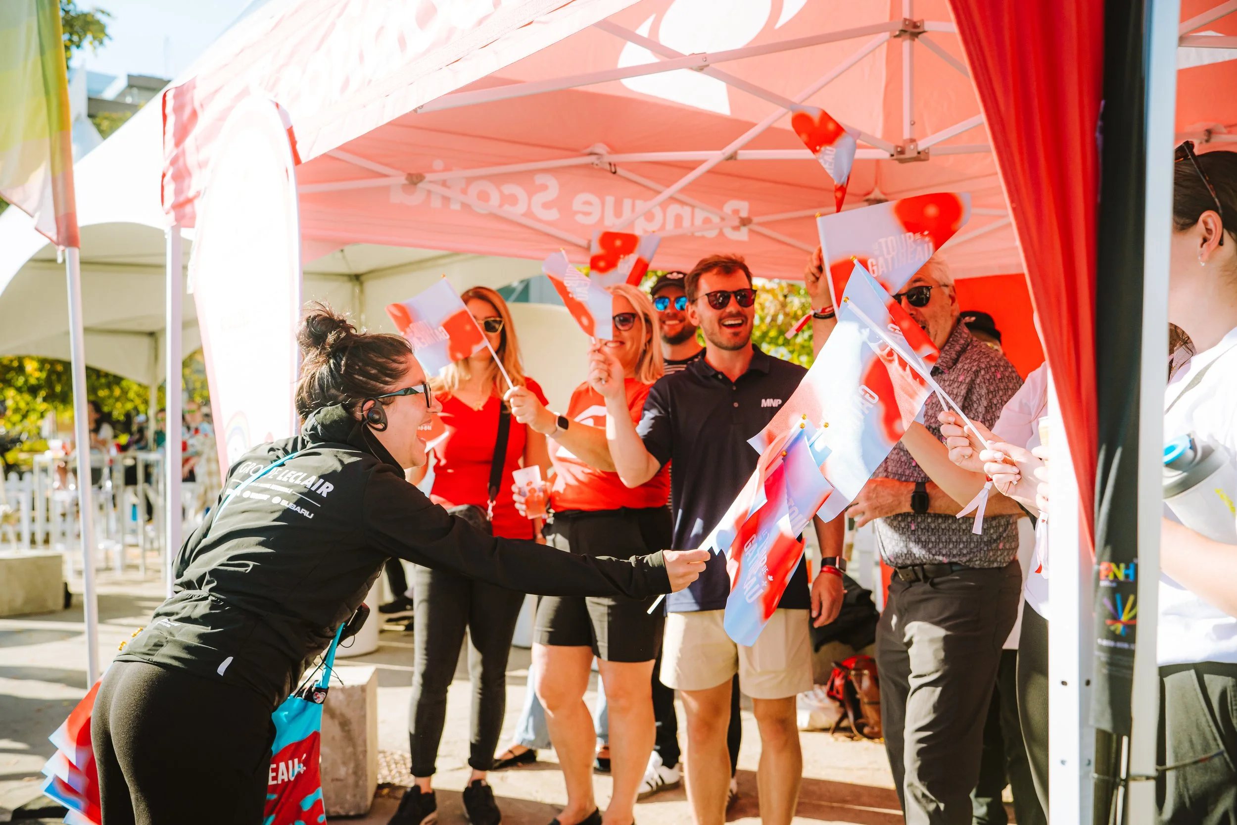 Group of cheerful people at an outdoor event under a pink canopy, waving flags and engaging with each other.