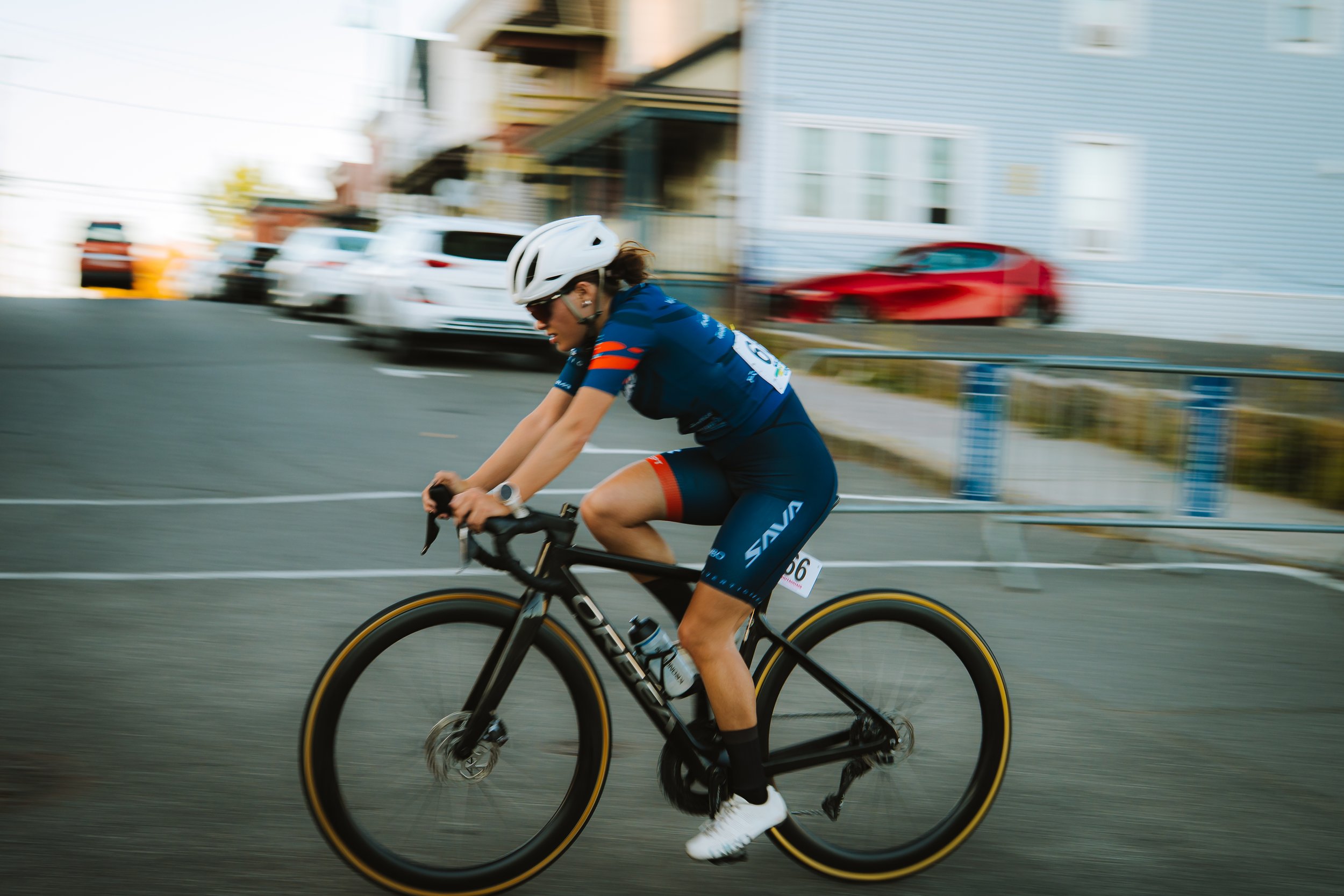 A person riding a black road bike during a race, wearing a blue cycling uniform, a white helmet, and sunglasses. The background shows blurred cars and houses, indicating motion.