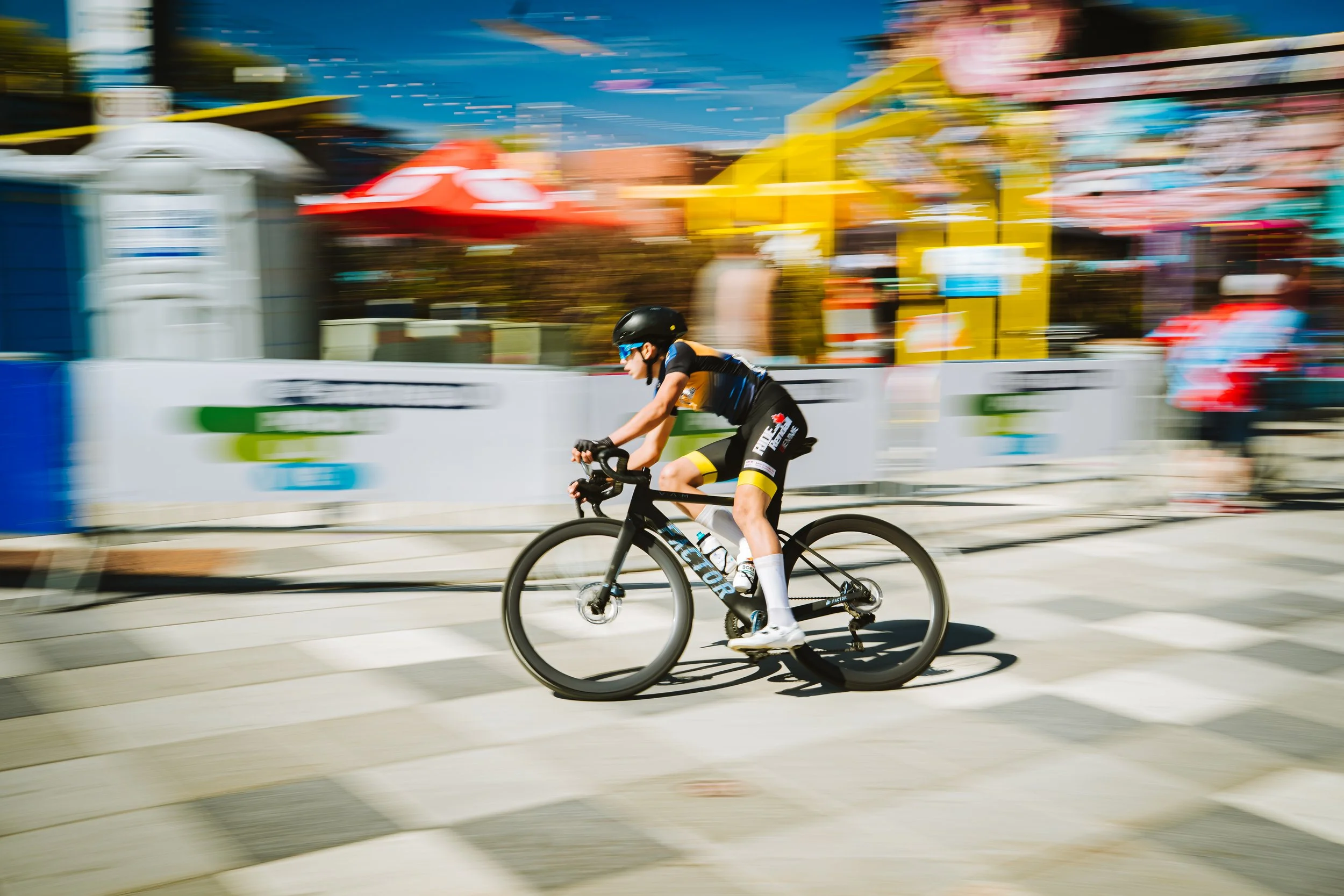 A cyclist riding a black bike at a high speed in an outdoor event with colorful amusement park rides and advertisements in the background during daytime.