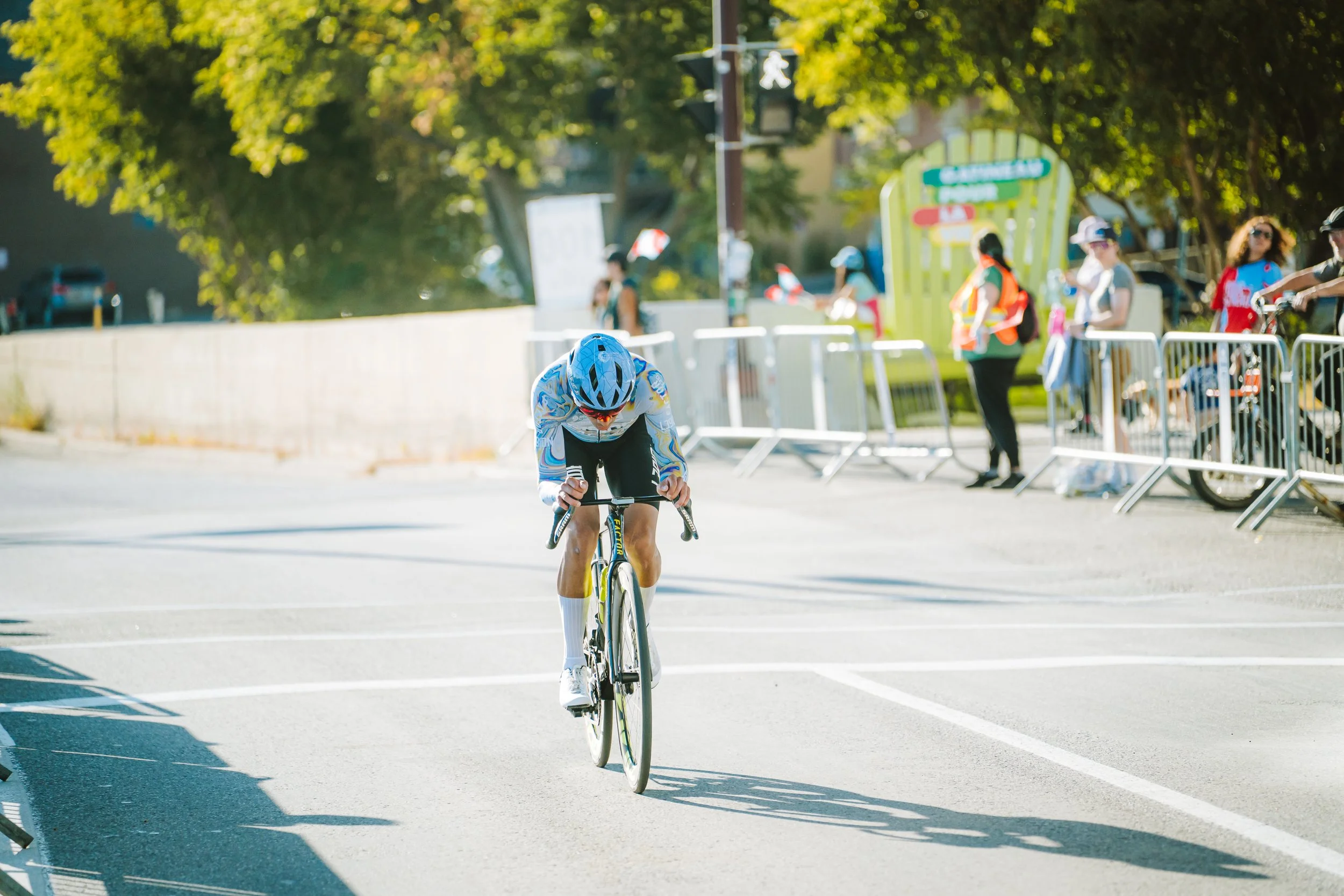 A cyclist wearing a blue helmet and white and blue cycling gear riding on a road during a race, with spectators behind barricades on the side of the street.