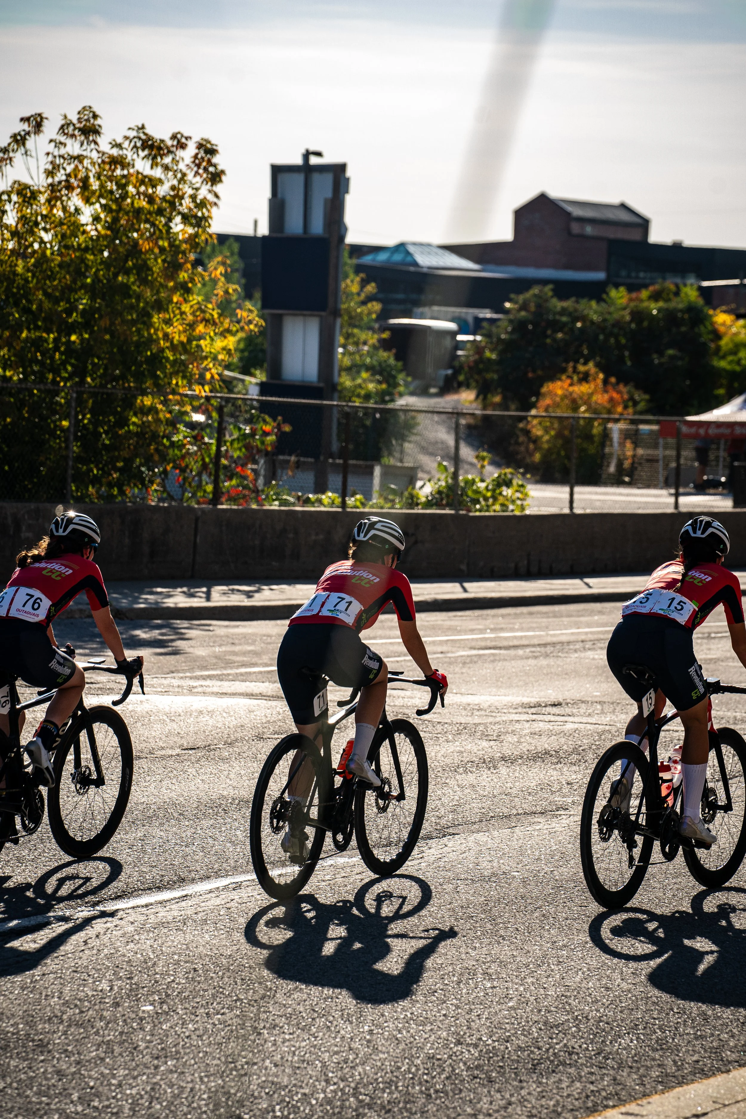 Three young cyclists in racing gear riding on a city street during a race, with sunlight casting their shadows on the road and trees and buildings in the background.