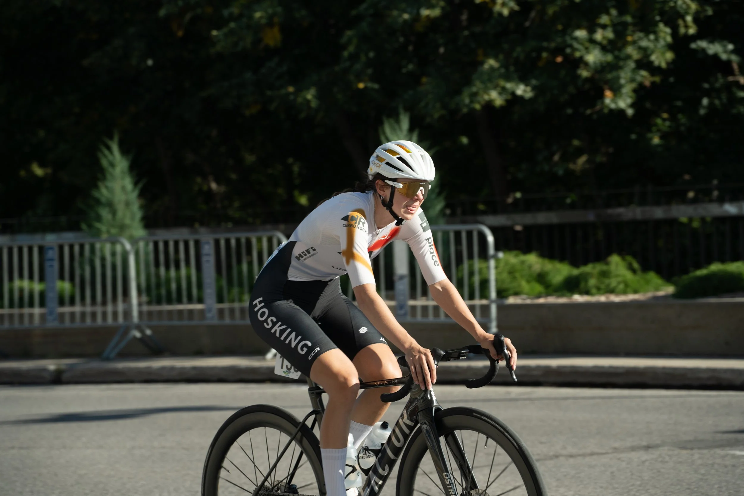 A female cyclist in a white and black cycling outfit, wearing a white helmet and sunglasses, riding a black road bike on a city street with trees and a metal barrier in the background.