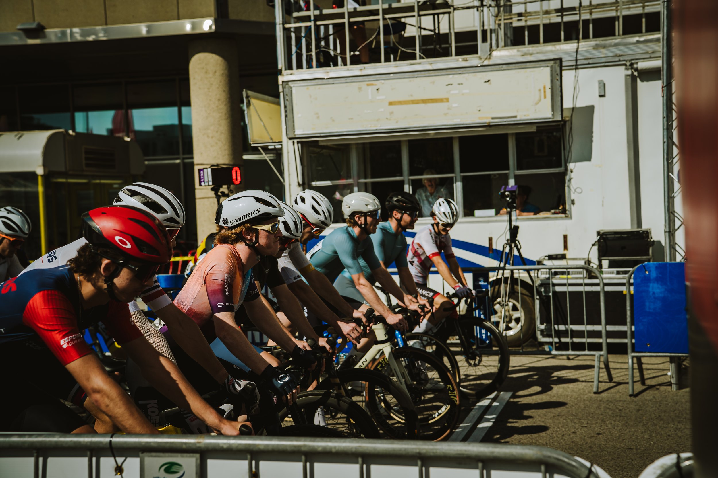 Cyclists at the start line of a race, lined up and ready to begin, wearing helmets and athletic gear, with a support vehicle and equipment in the background.