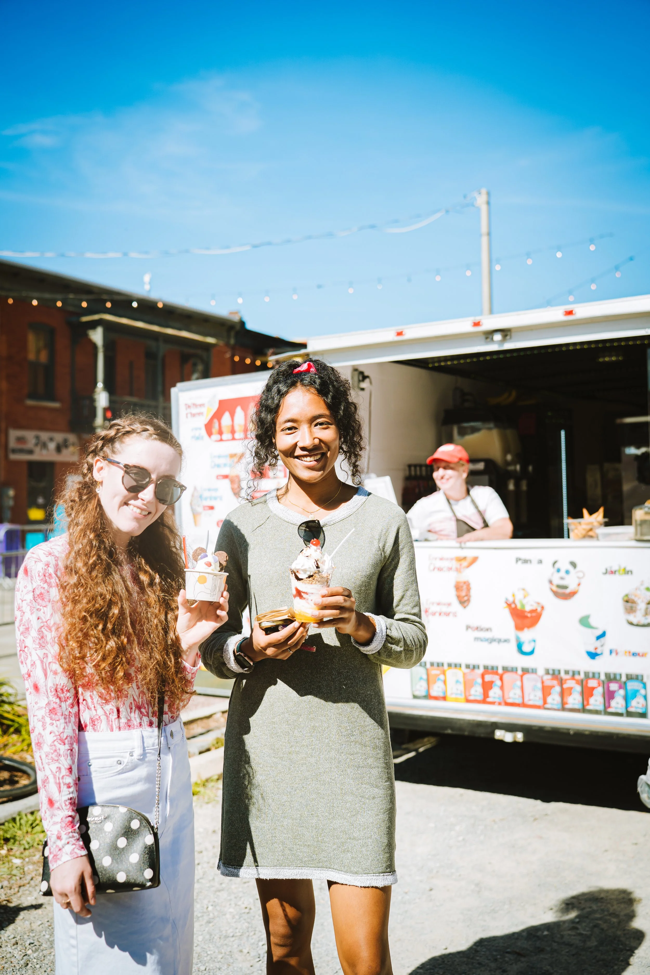 Two women smiling and holding ice cream cones at an outdoor event with a food truck in the background.