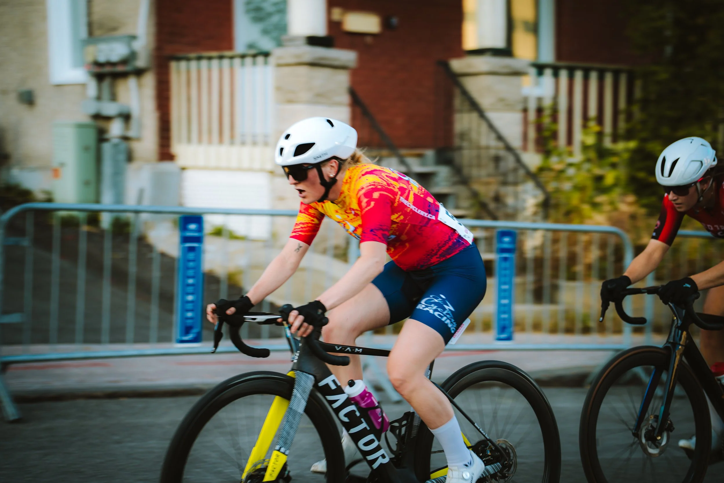 Female cyclist in a red and yellow jersey riding a yellow and black bike during a race, wearing a white helmet and sunglasses, with another cyclist in a red jersey nearby.