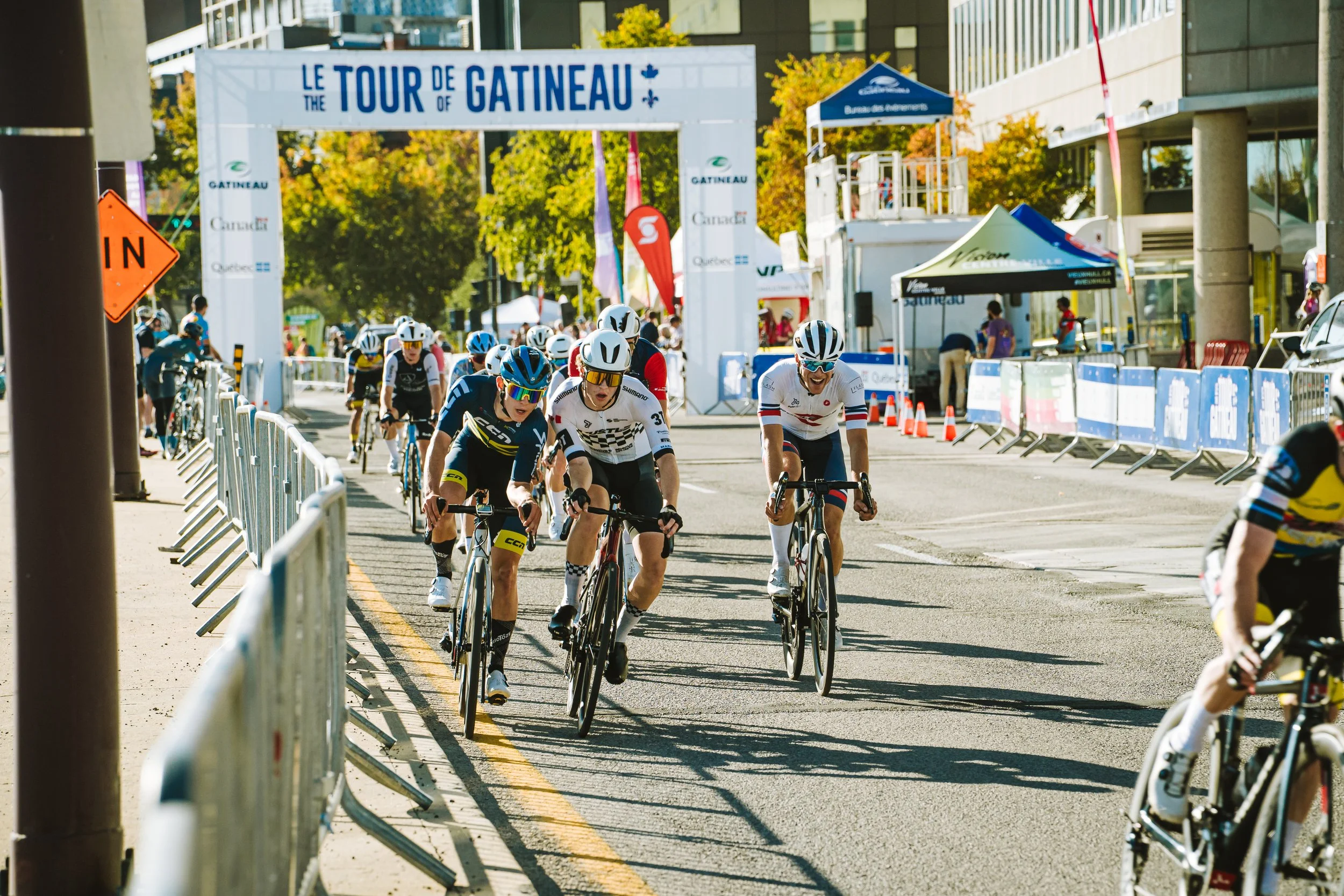 Cyclists racing on a city street during the Tour de Gatinau event, with banners, barricades, and spectators in the background on a sunny day.