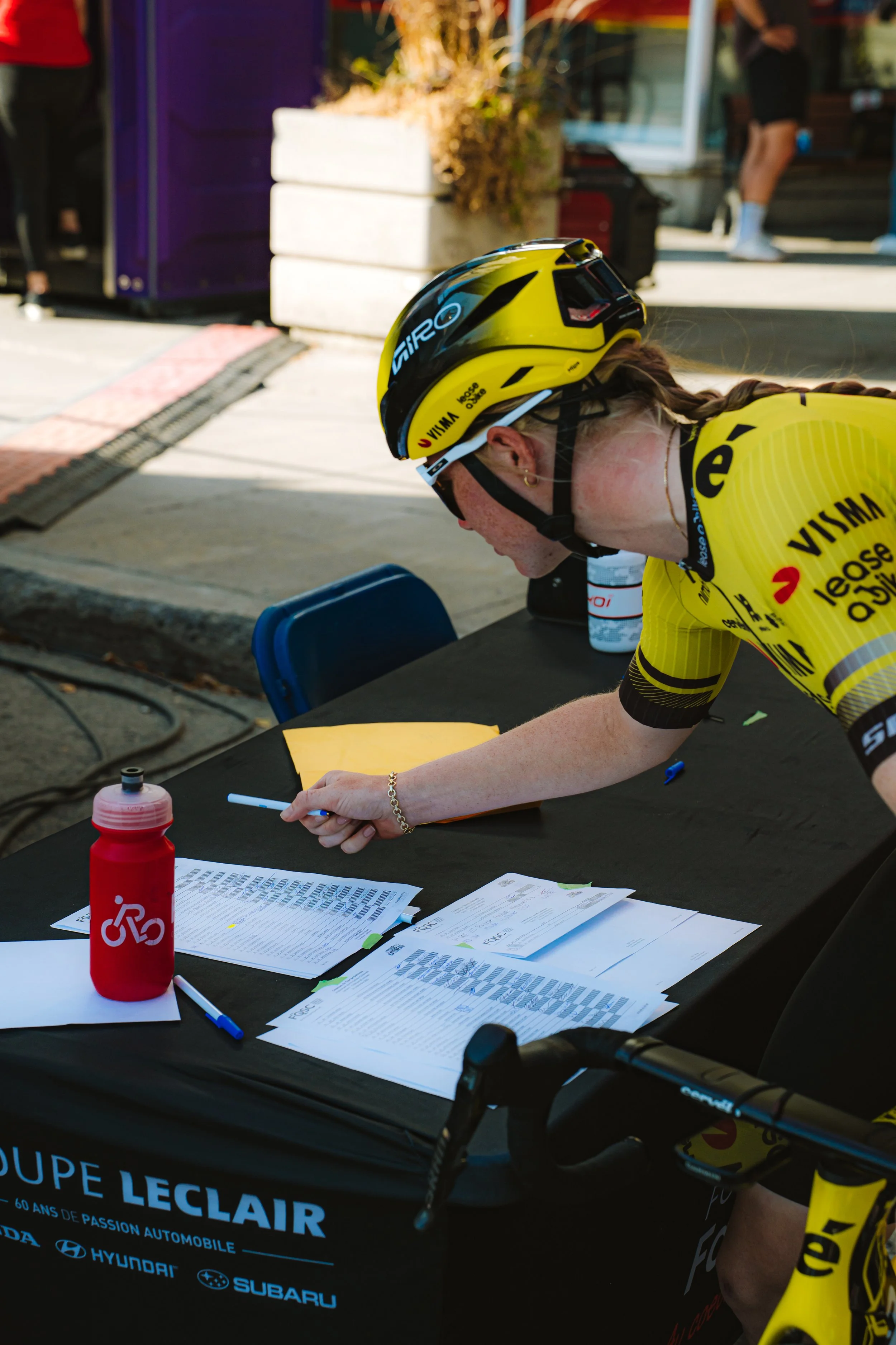 A female cyclist wearing a yellow team jersey and helmet is signing documents on a black table outdoors during daytime. There are papers, a red water bottle, and a blue pen on the table.