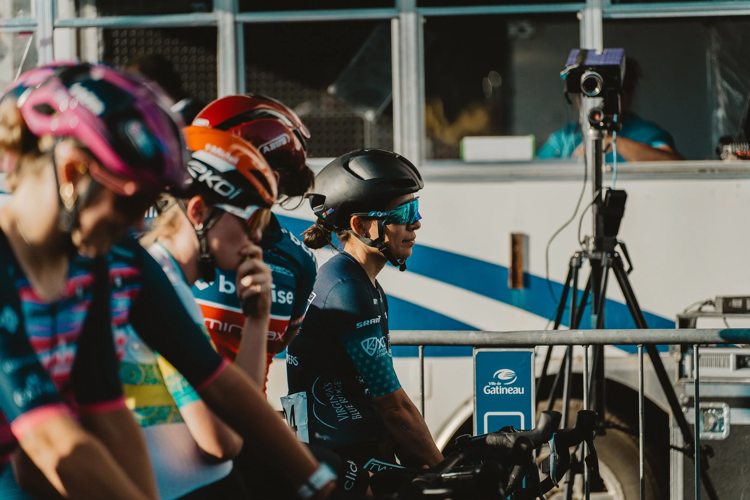 Group of female cyclists wearing colorful helmets and jerseys, preparing at the starting line of a race with a support vehicle and camera setup in the background.