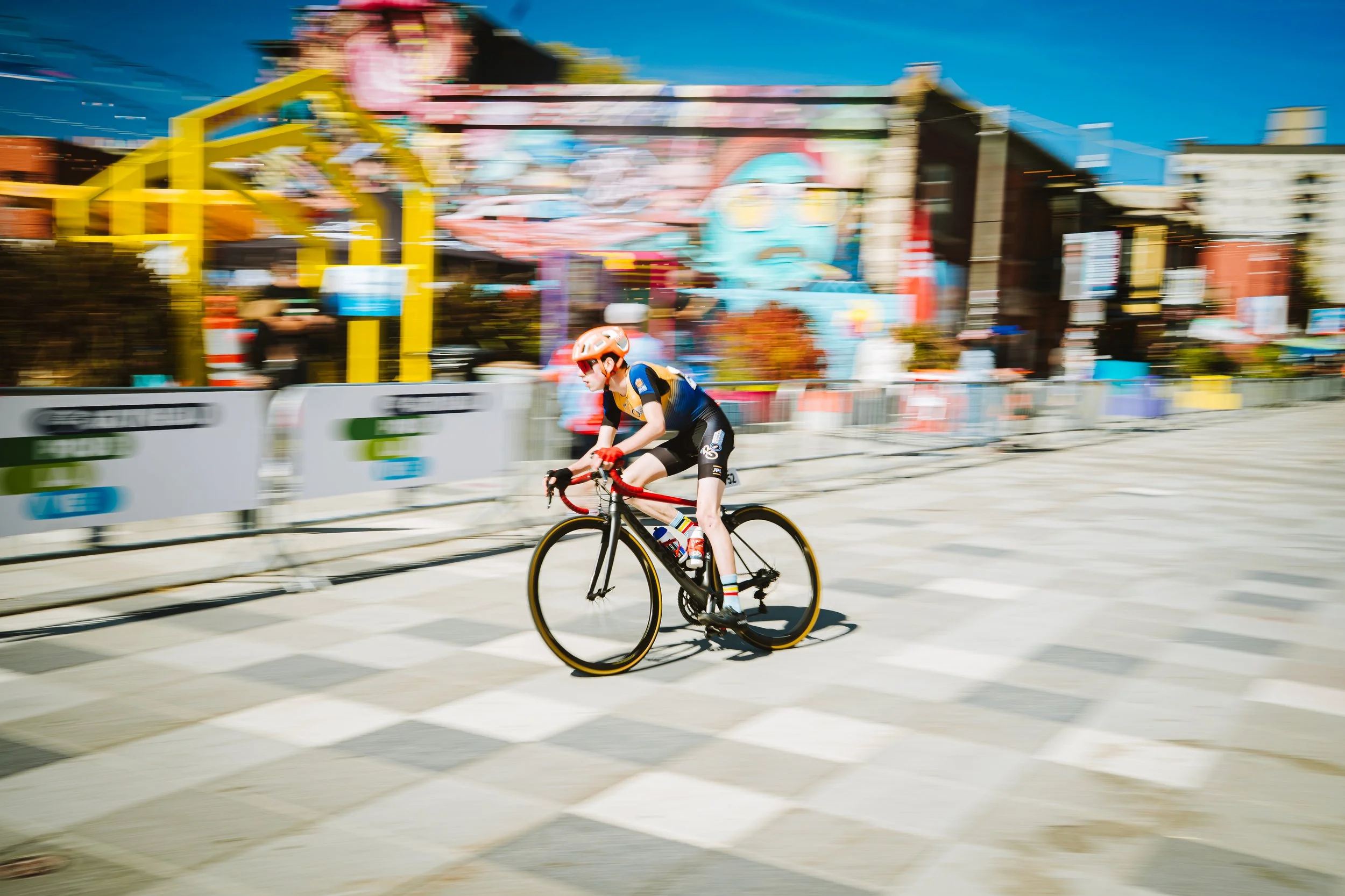A cyclist wearing a helmet and sports attire riding a bike on a city street with colorful background and blurred surroundings.