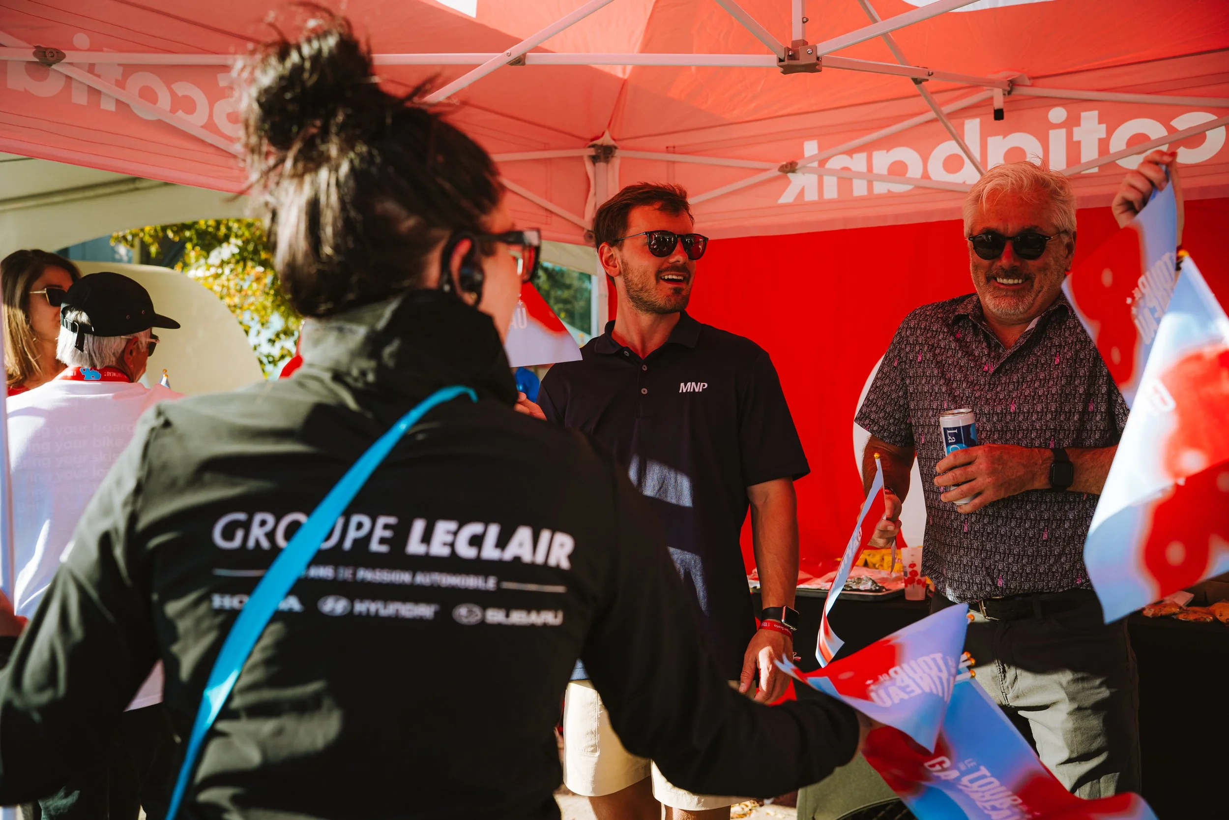 People gathered under a red canopy, smiling and talking. One person is wearing a jacket with 'GROUPE LECLAI' written on it. Two men are holding flags and a can of beverage, smiling and with sunglasses. The scene appears to be a casual outdoor event w