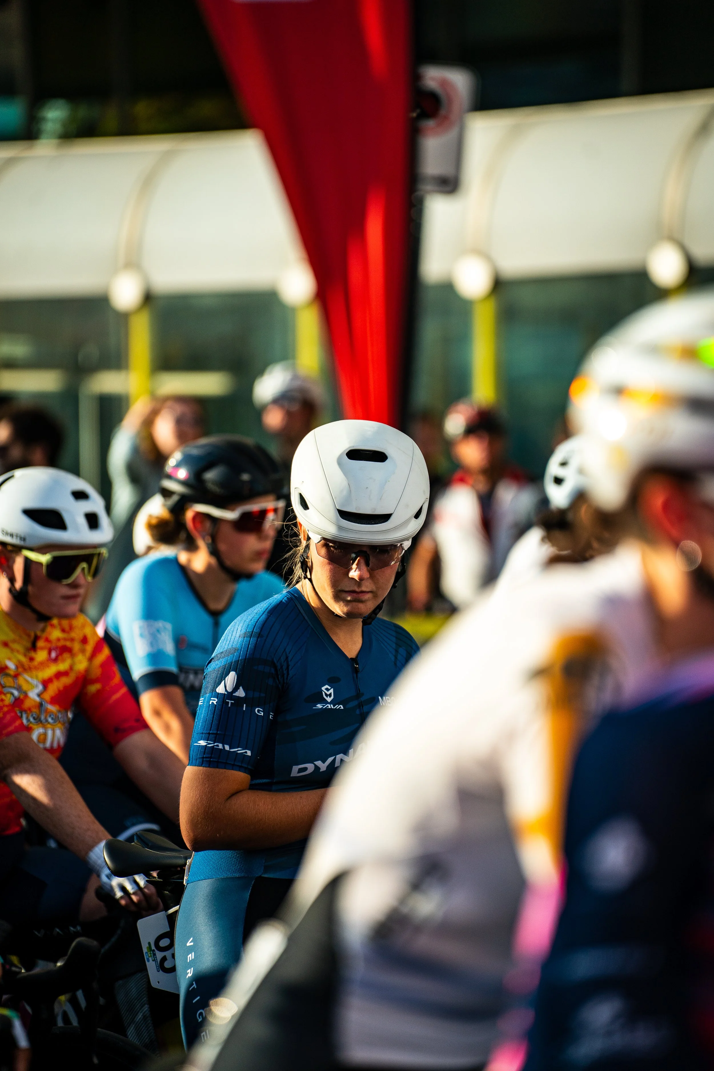 Group of female cyclists wearing helmets and sunglasses at the starting line or beginning of a race, with some focusing intently and others looking around, under a red canopy.