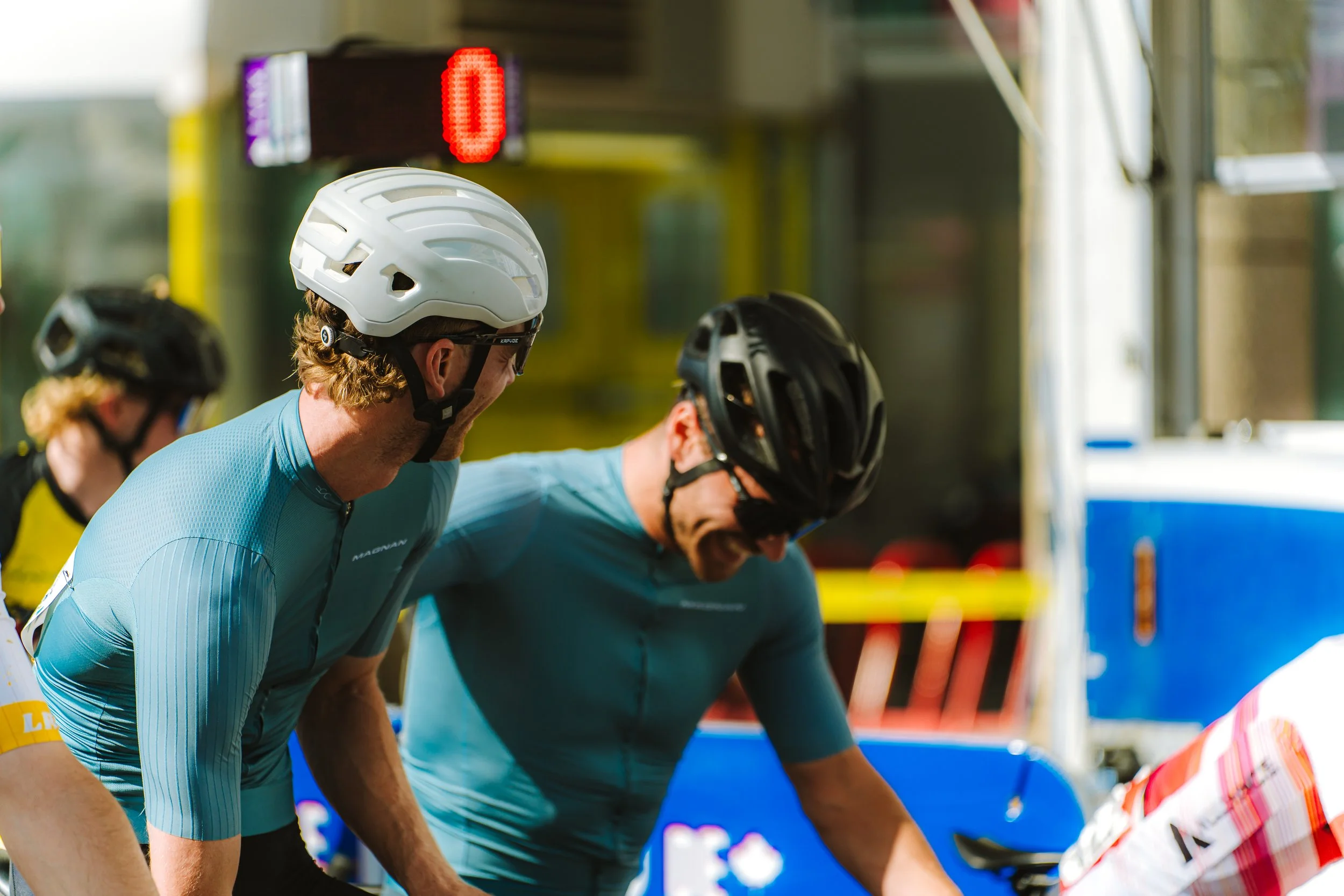 Cyclists in racing gear wearing helmets, bending over their bikes, at a race start or finish line.