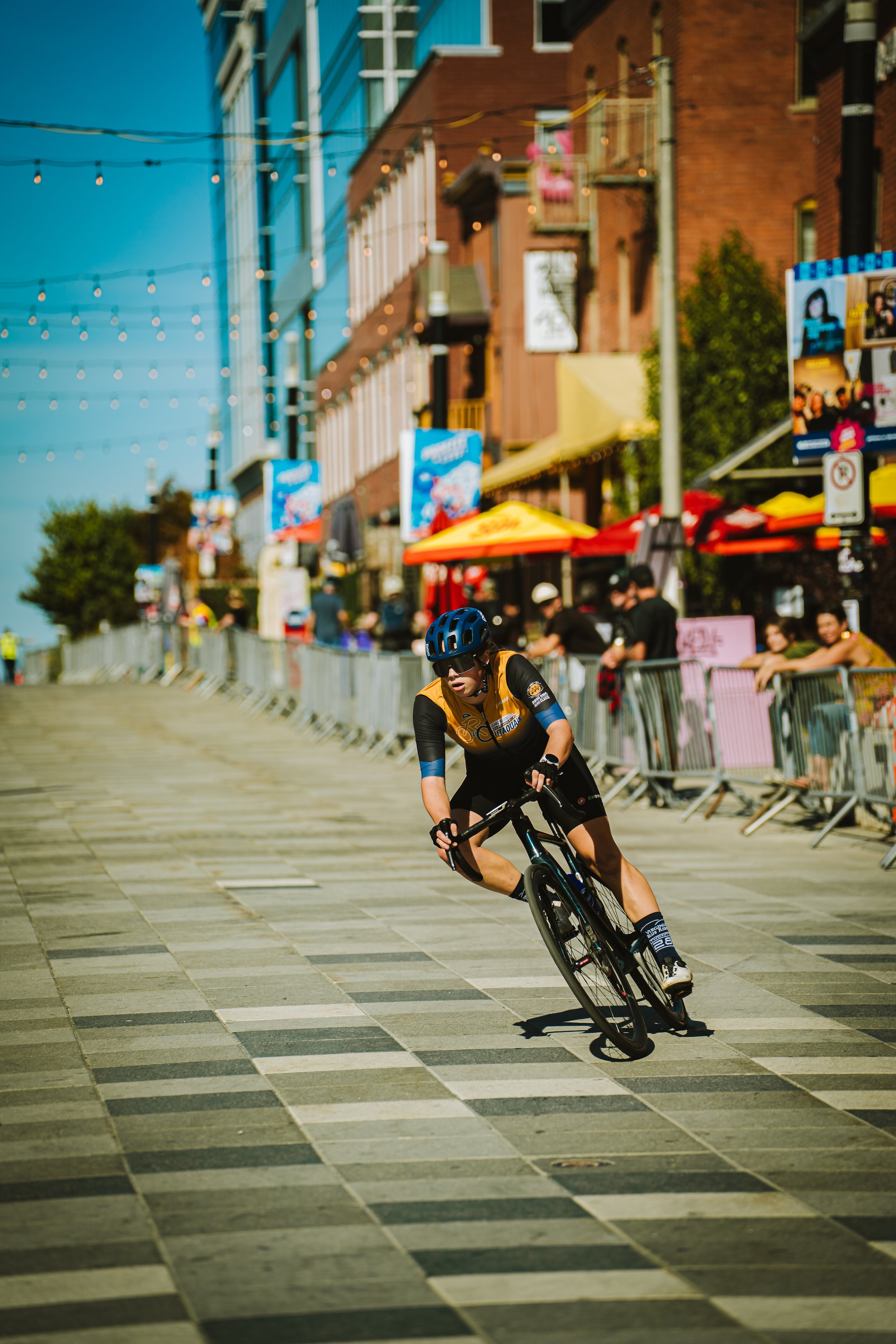 Cyclist wearing a blue helmet and black sports gear rides on a city street with barriers and spectators on the side, colorful storefronts, and buildings in the background.