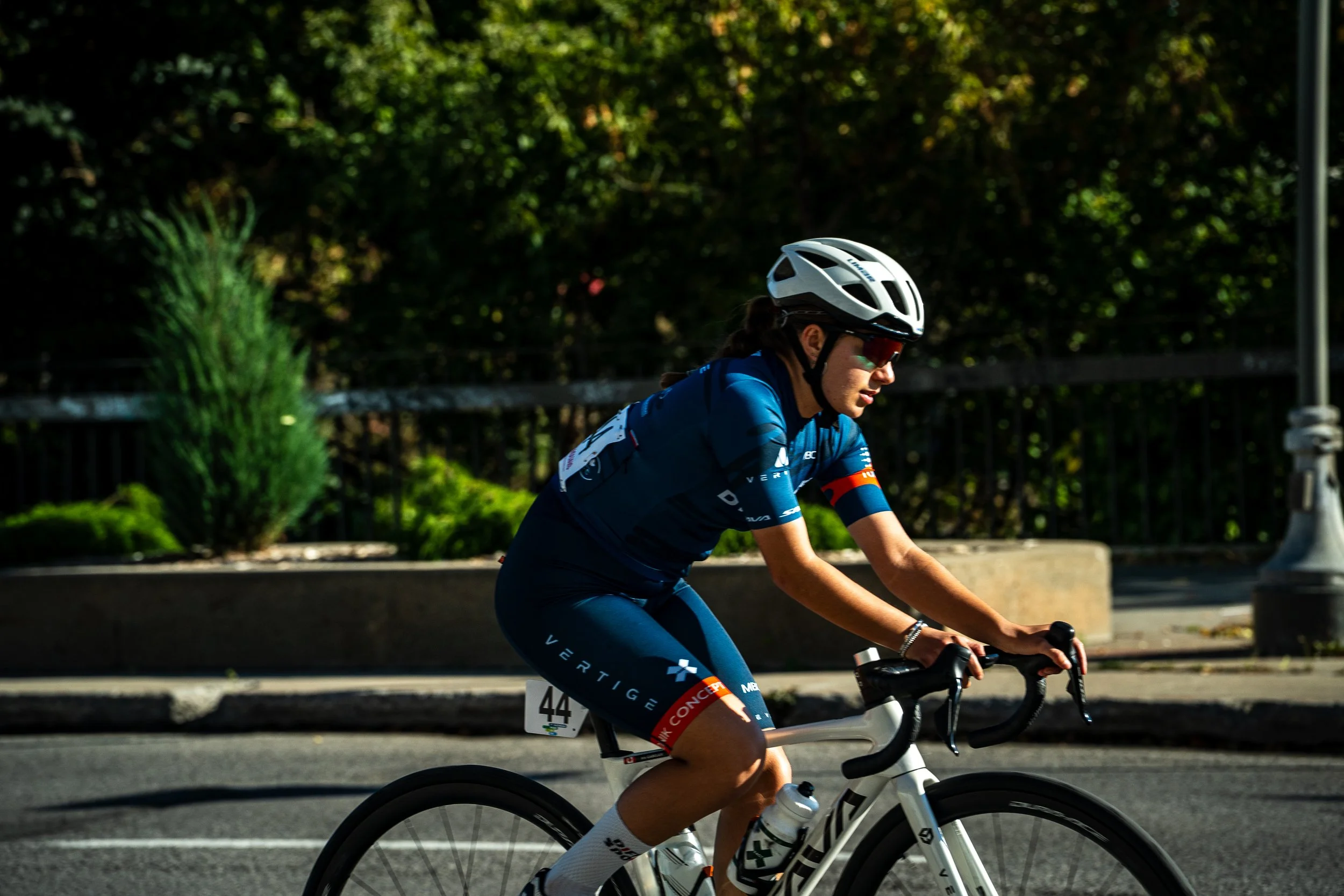 Female cyclist wearing a blue jersey, dark blue shorts, white helmet, and sunglasses riding a white racing bike on a road with greenery in the background.