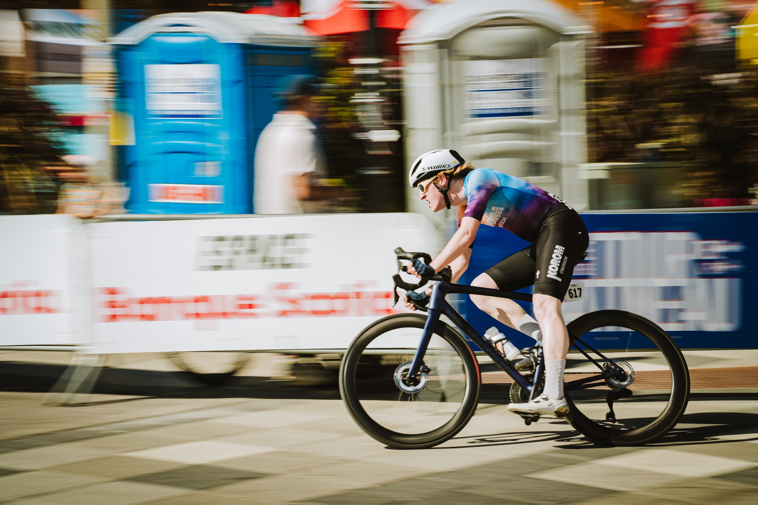 Cyclist racing on a bike in a competitive event with spectators and colorful barriers in the background.