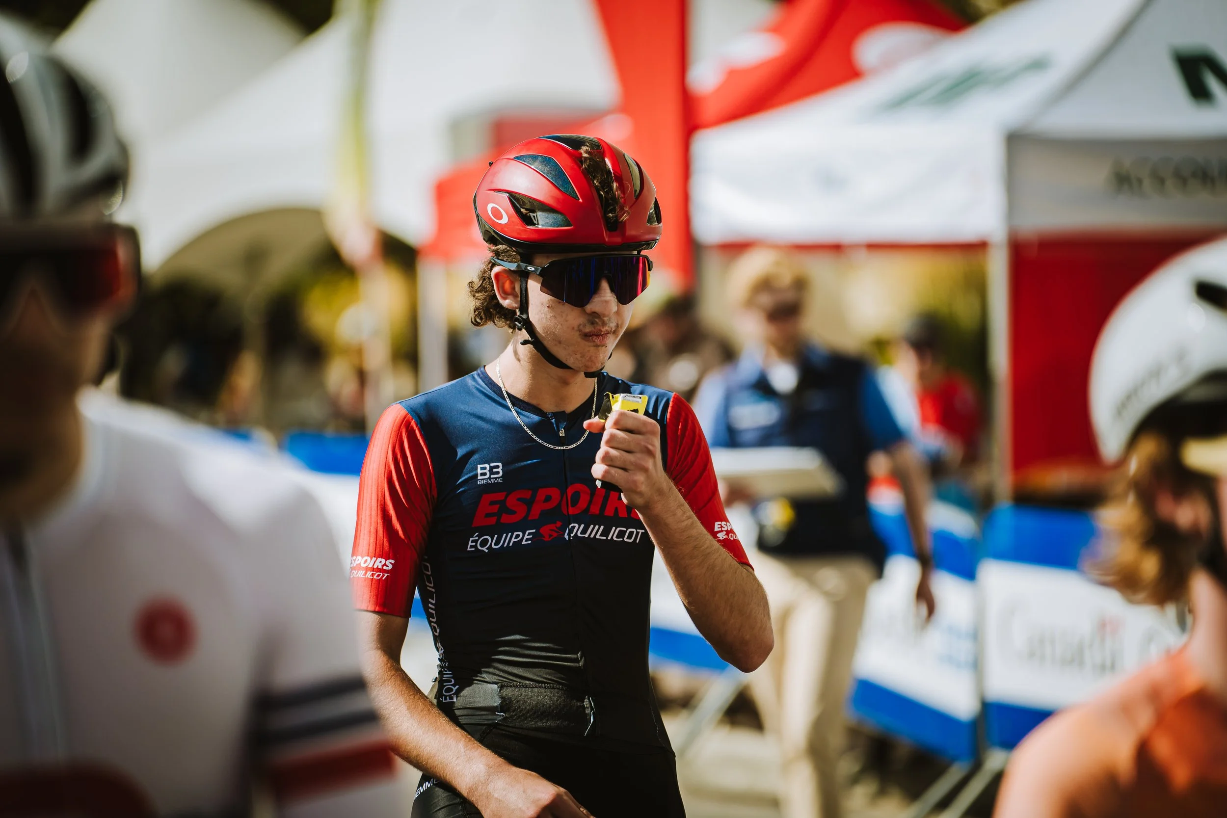 A young man in a red and black cycling jersey, wearing a red helmet and dark sunglasses, looks at a device in his hand, likely at a cycling event.