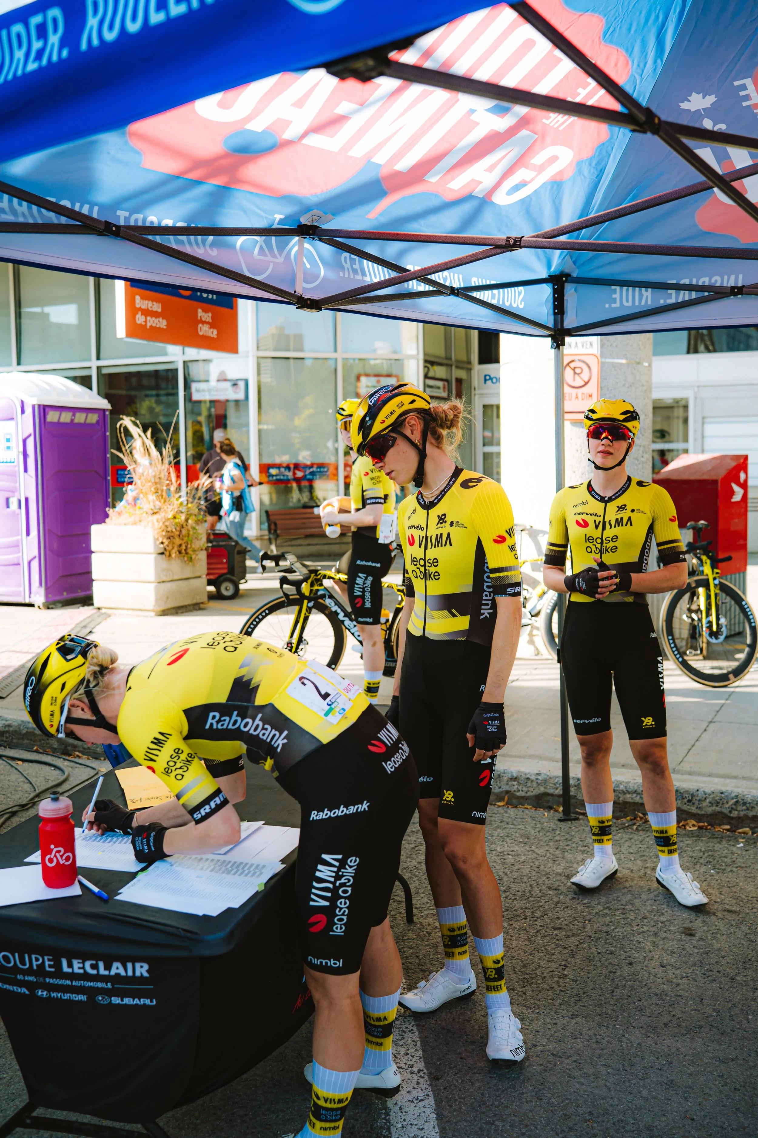 Cyclists in yellow and black team uniforms preparing at a registration table beneath a blue canopy at a cycling event.