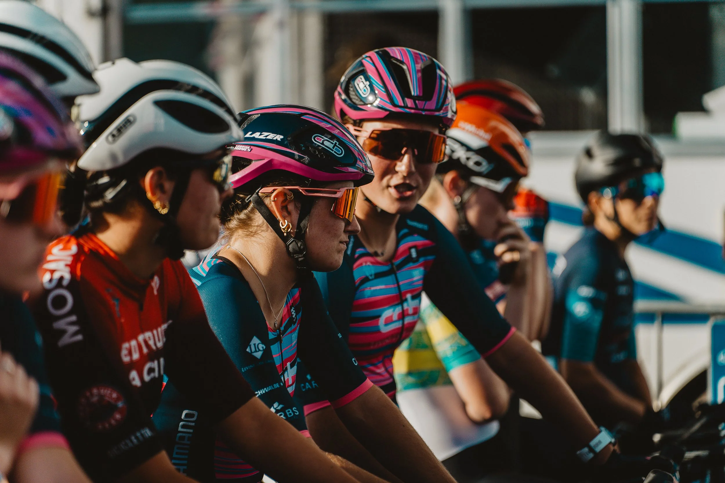 Group of female cyclists wearing helmets and sunglasses preparing for a race.