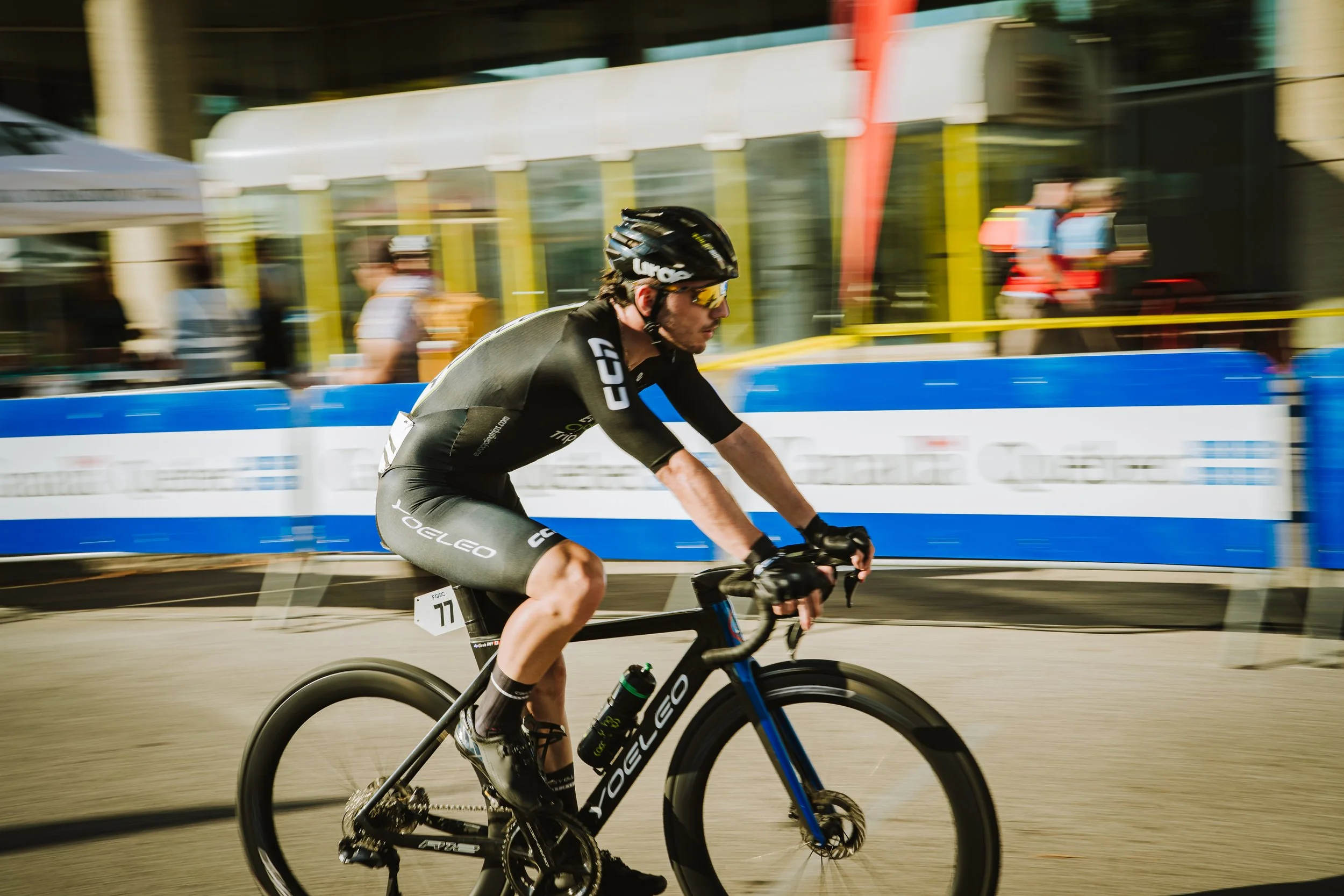 A male cyclist racing during a daytime event, wearing a black helmet, sunglasses, and a black and gray cycling outfit, riding a black and blue road bike. The background is blurred with cityscape elements and event barriers, indicating motion and spee