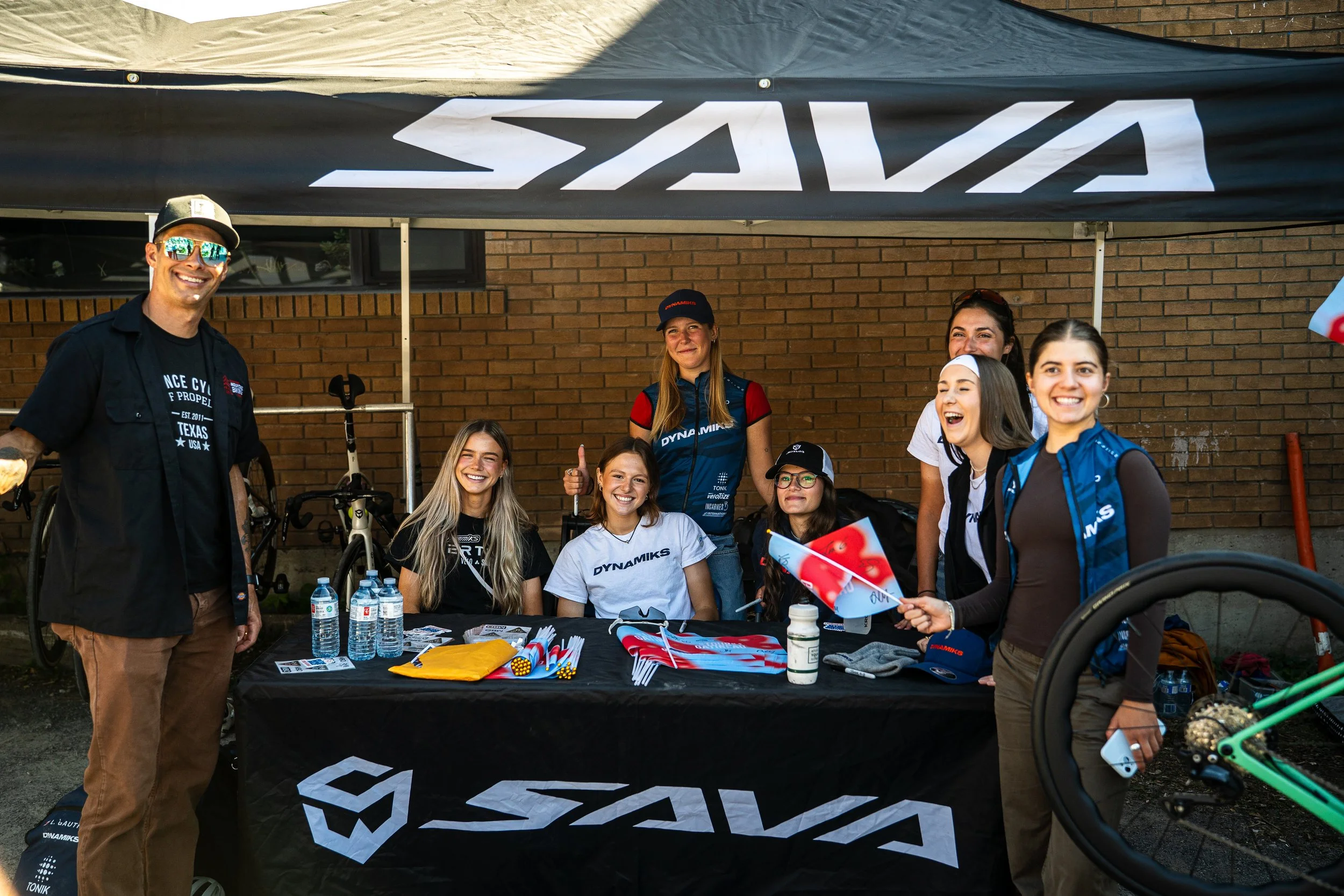 Group of smiling women and a man under a black canopy with the 'SAVA' logo, sitting behind a table with merchandise and water bottles, in front of a brick wall. One woman holds a small American flag.