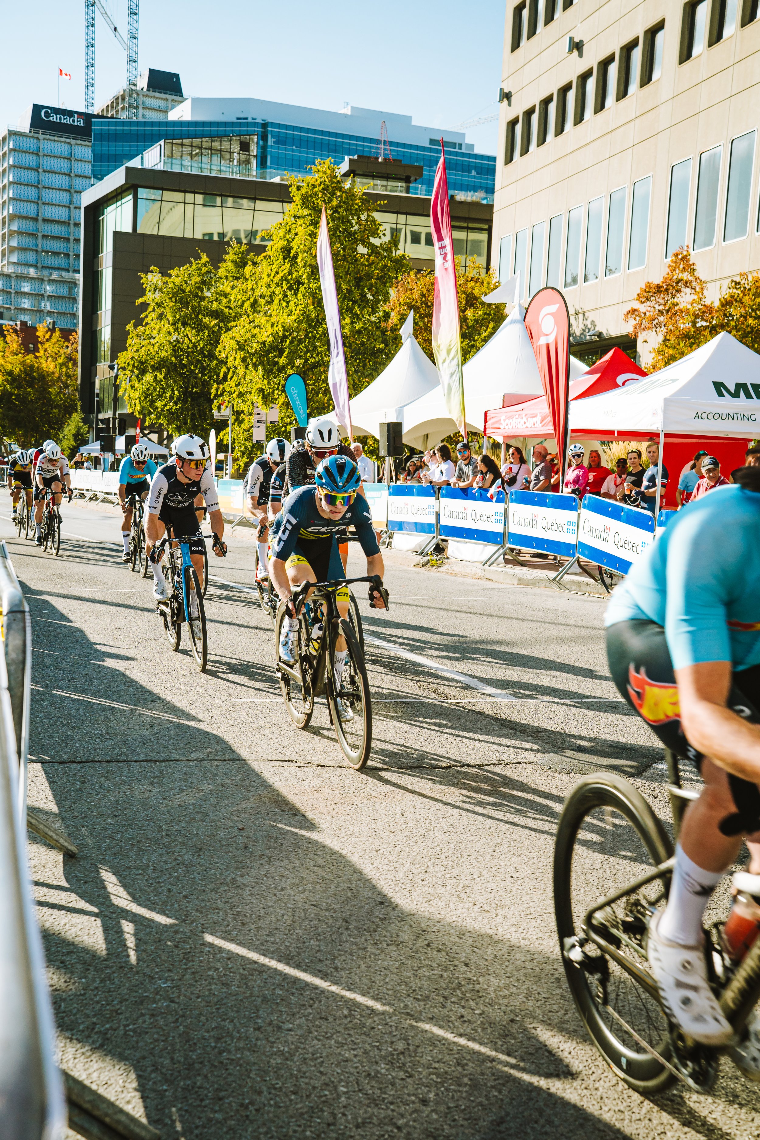 Cyclists participating in a city street race with spectators and tents on the sidewalk, tall buildings in the background, and bright sunny weather.