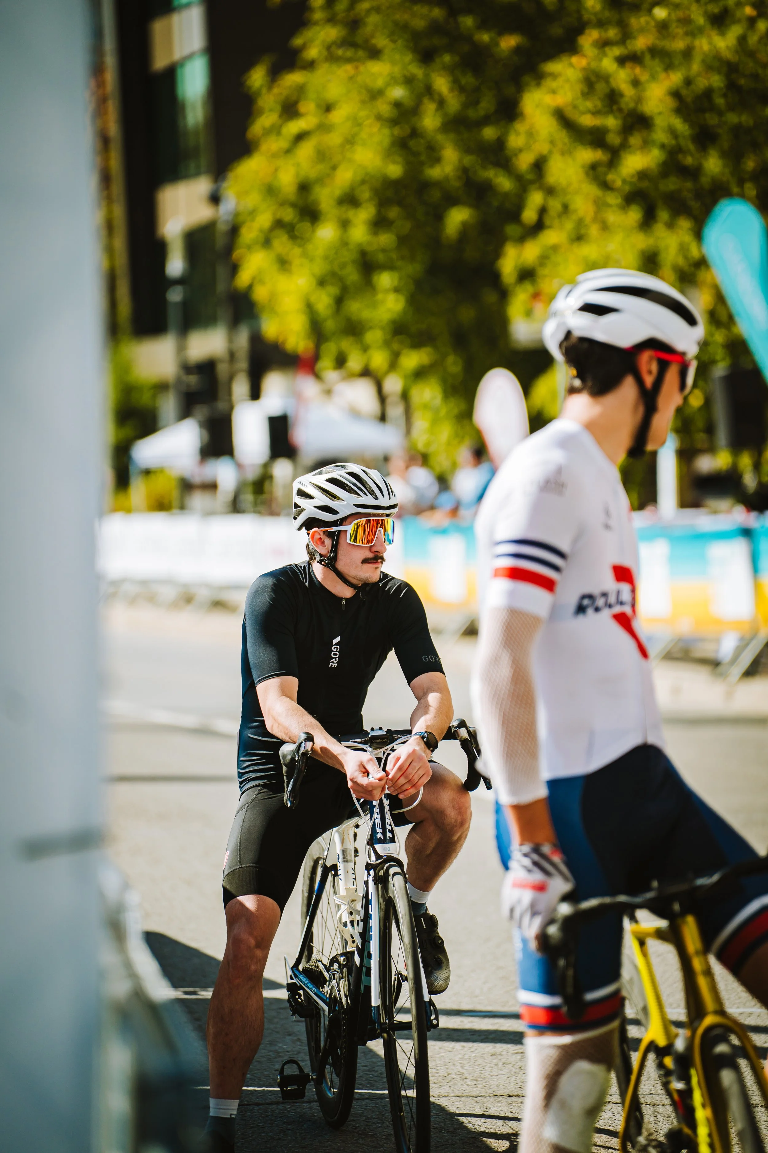 Two male cyclists wearing helmets and sunglasses, one sitting on a black bicycle and the other standing beside a yellow bicycle, at an outdoor event on a sunny day with trees and tents in the background.