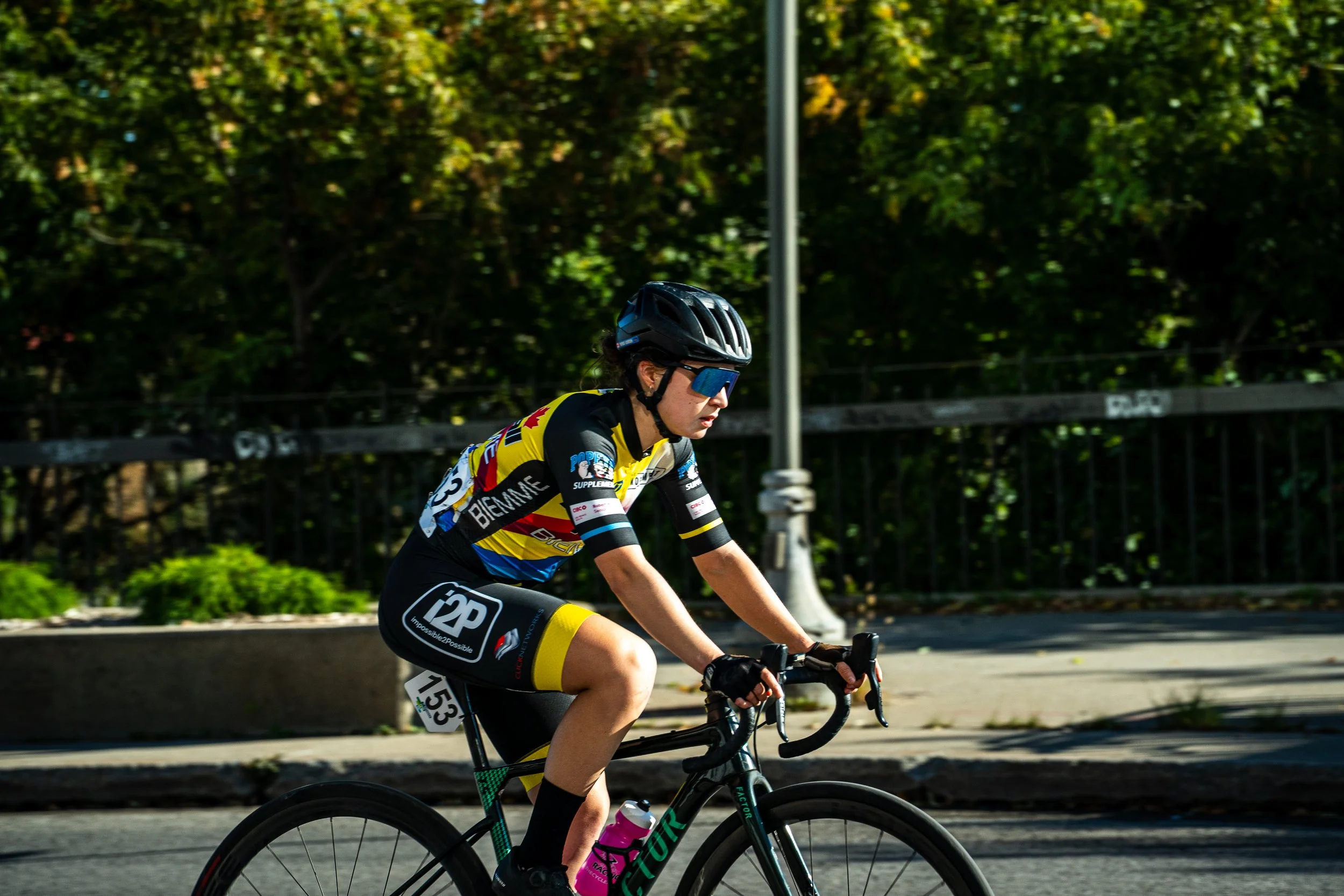 Female cyclist in black, yellow, and blue cycling jersey and black shorts riding a road bike with a pink water bottle.