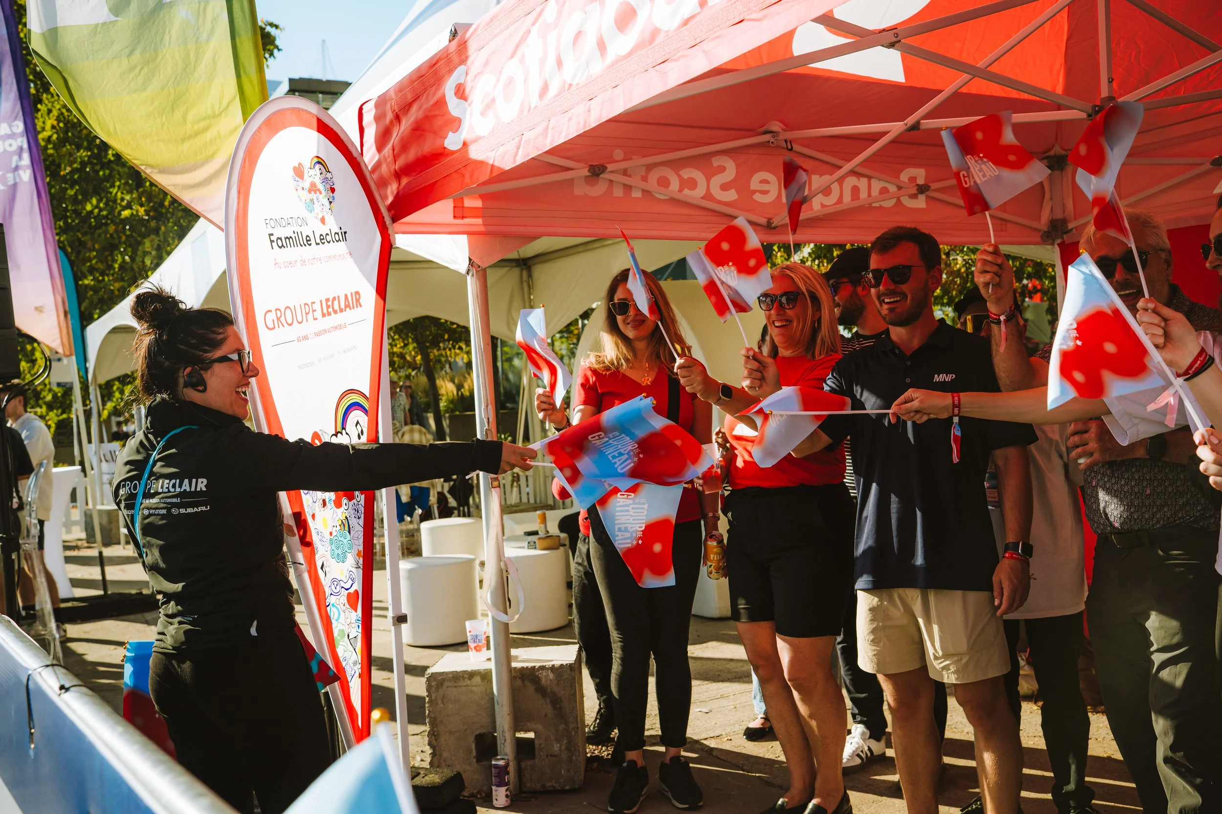 Group of people at an outdoor event under a red tent, waving small Quebec flags. Some are wearing sunglasses and smiling.