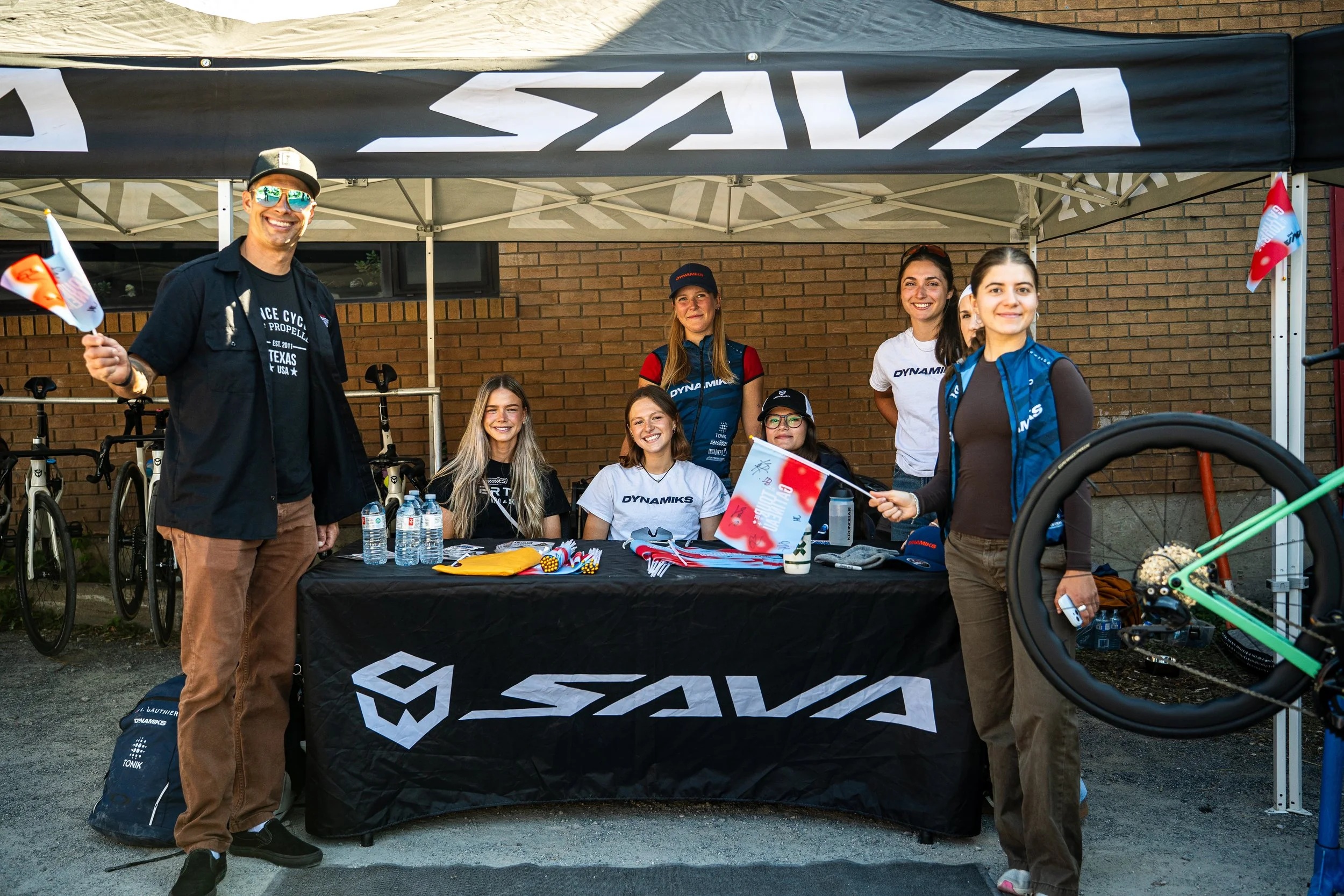 Group of people at an outdoor event under a black canopy with the 'SAVA' logo, sitting and standing behind tables with beer bottles, water bottles, and event merchandise, with a brick building in the background.