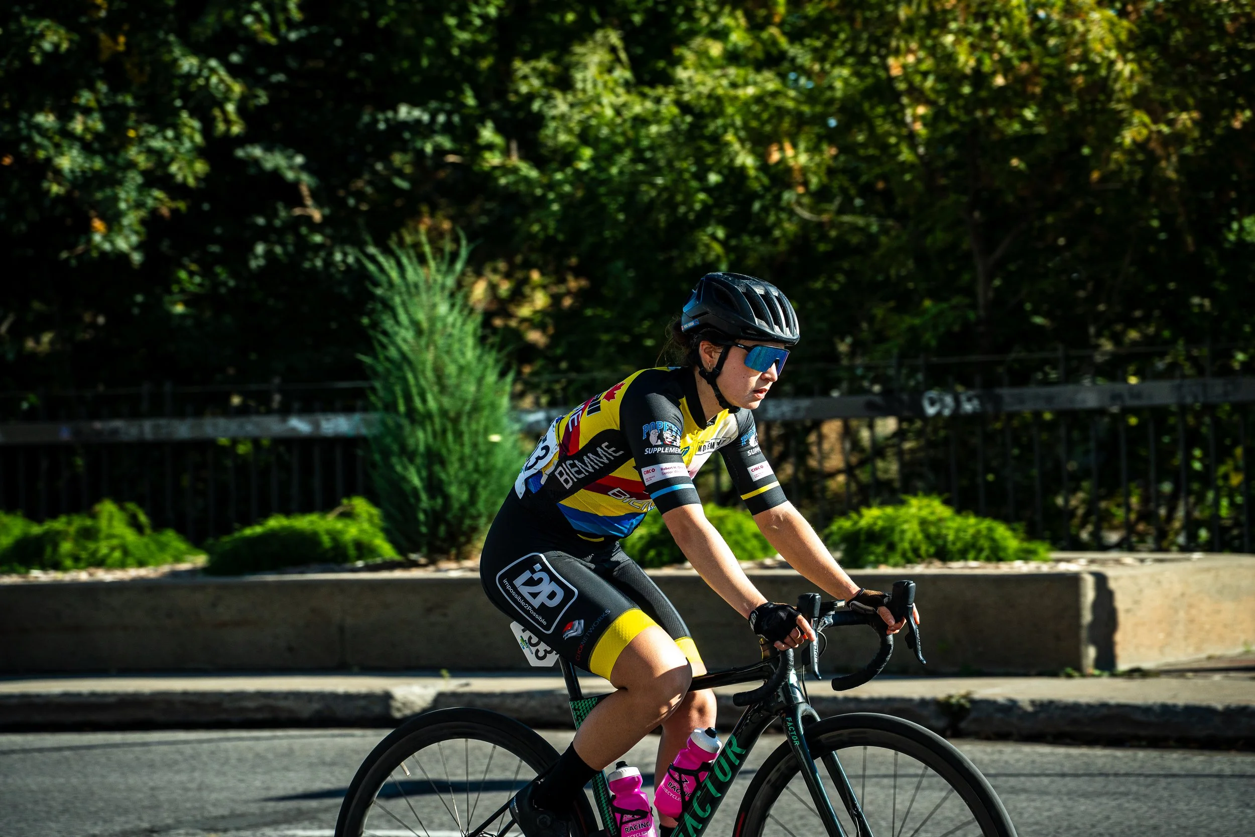A cyclist wearing a black helmet, blue sunglasses, and a black, yellow, and white cycling jersey riding a black bicycle on a street lined with green bushes and trees.