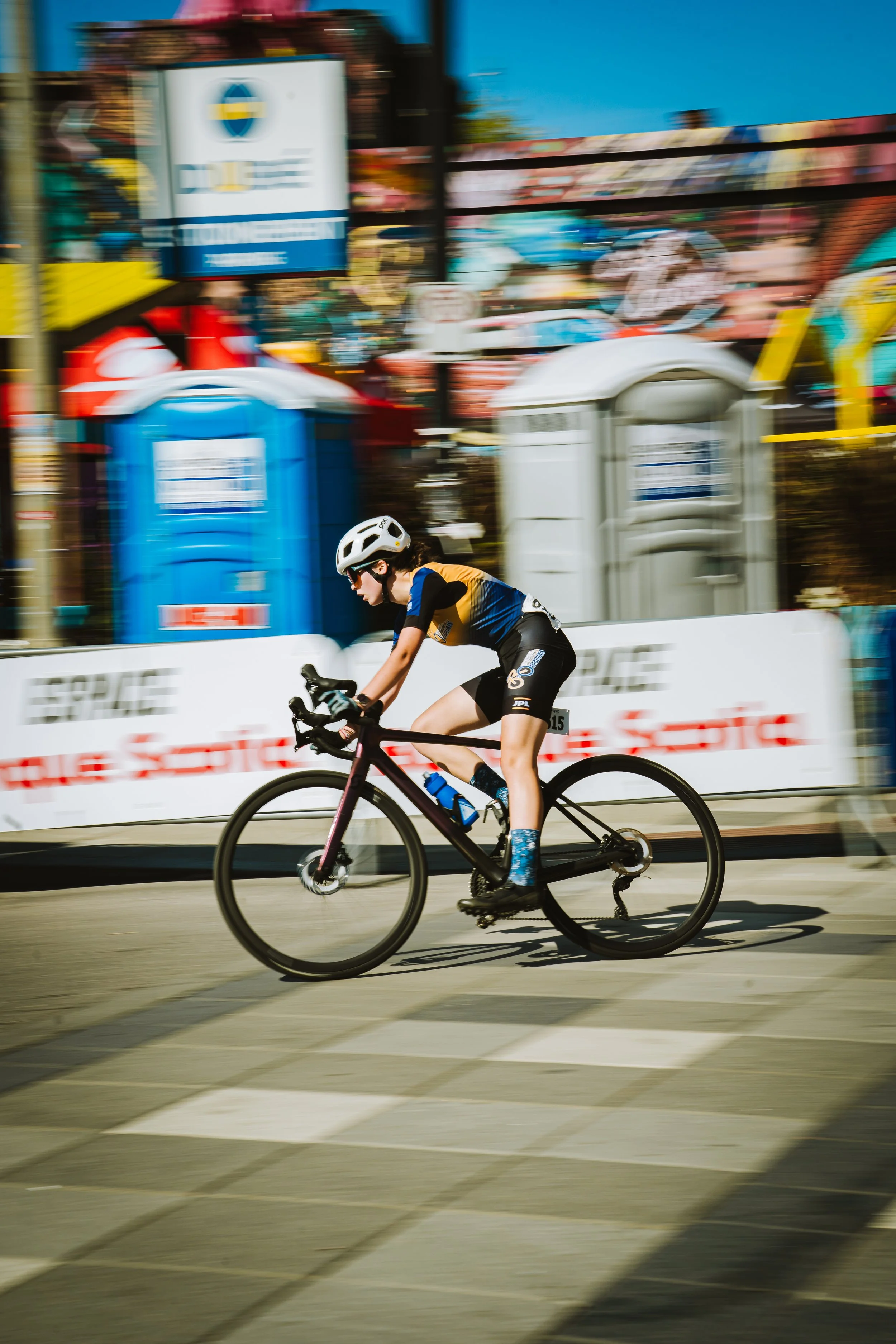 A cyclist wearing a white helmet, sunglasses, and a blue, black, and yellow jersey rides a black bike across a crosswalk during a race. The background is blurred with colorful banners, portable toilets, and a sign, creating a sense of speed.