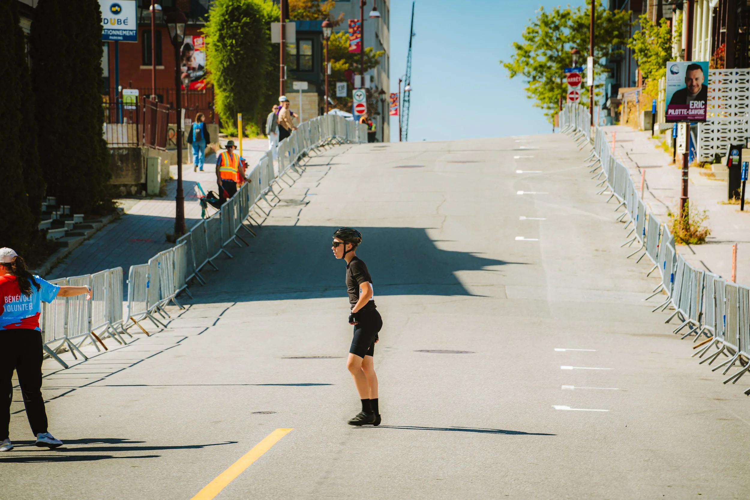 A woman in cycling gear and helmet standing on a closed town street with barricades, with people walking in the background and buildings on either side.