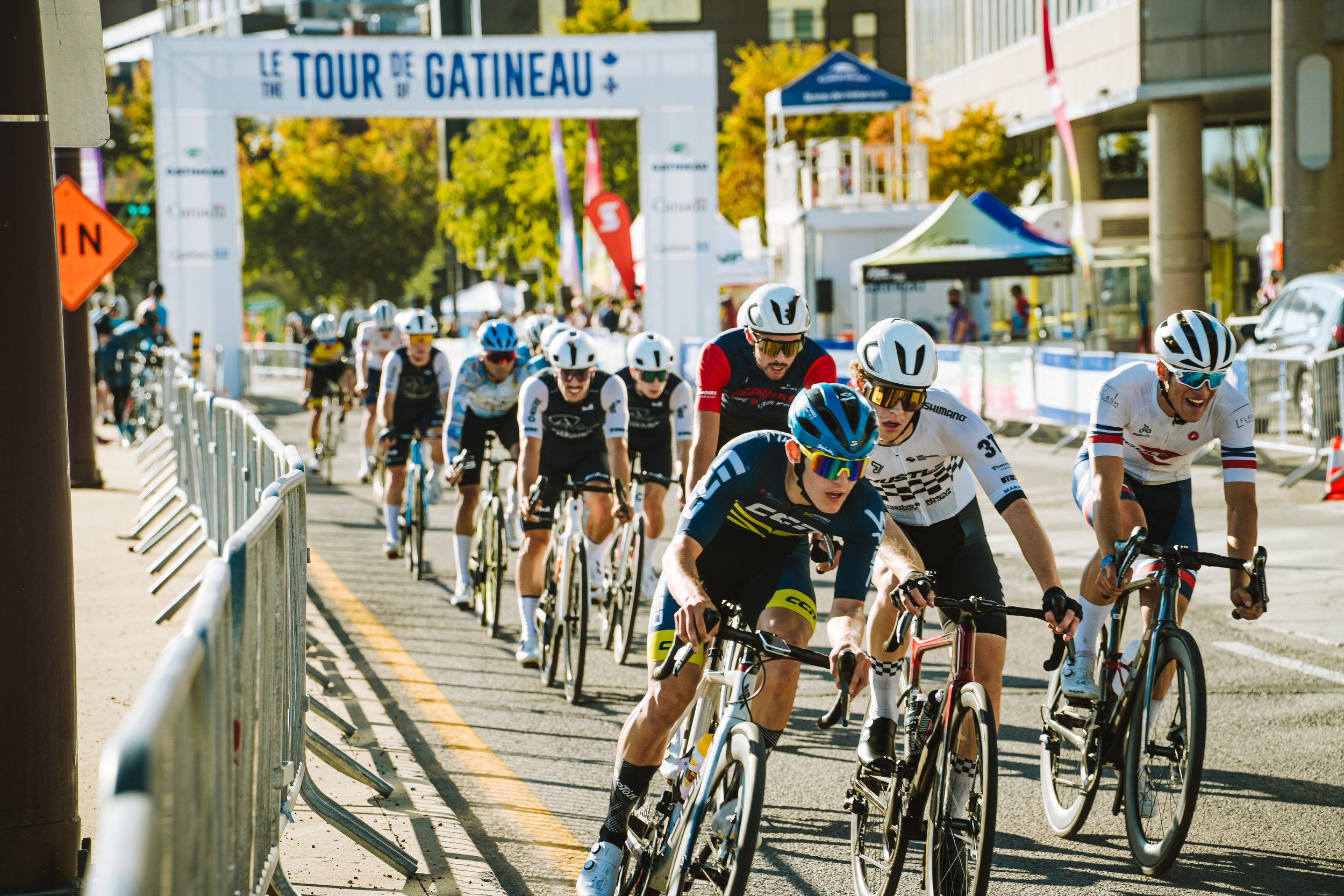 Group of cyclists participating in a race at the Tour de Gatineau event, riding on a city street with barriers and a large event banner overhead, with trees and buildings in the background.