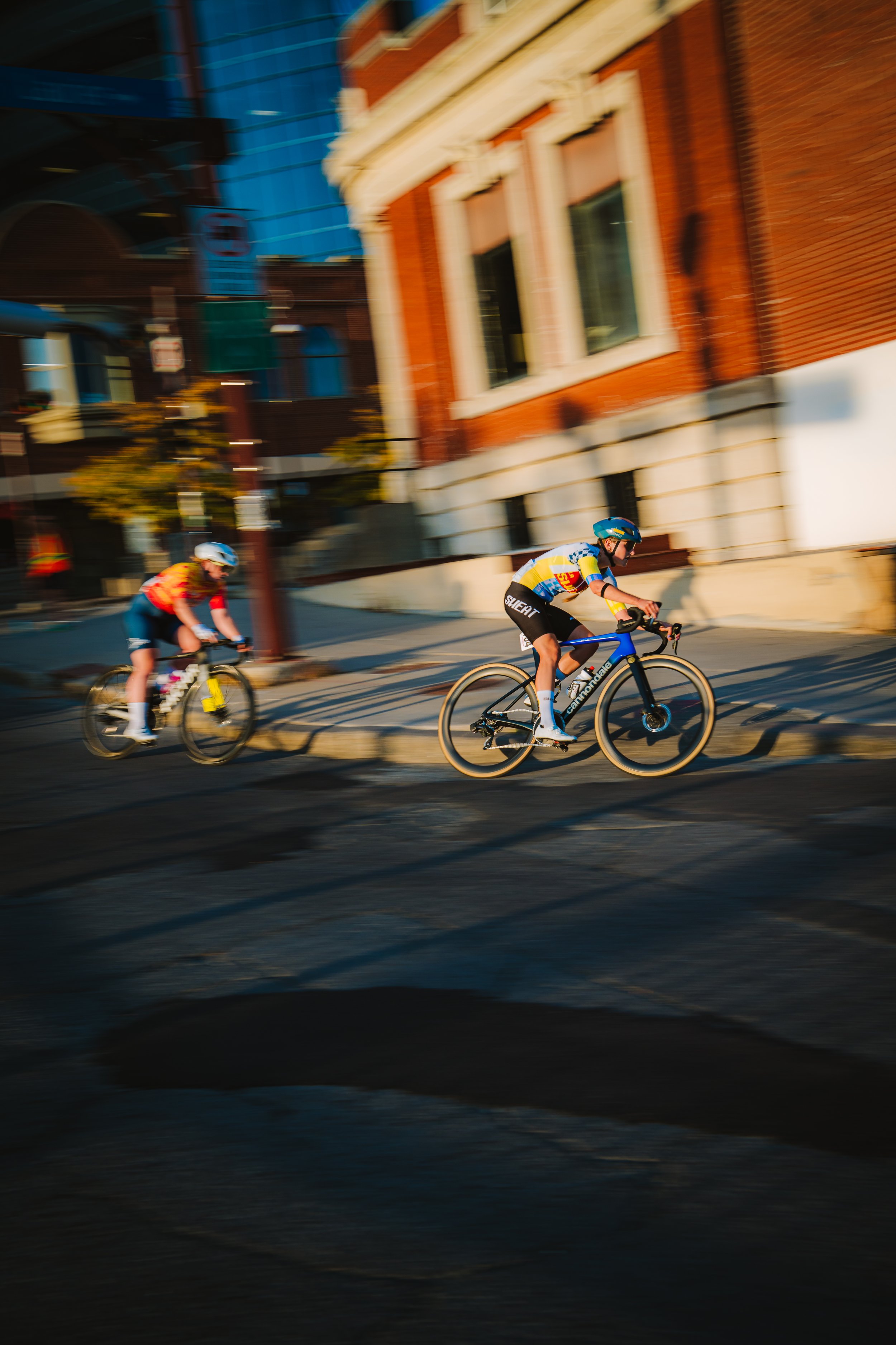 Two children riding bicycles on a city street during daytime, with blurred buildings in the background.