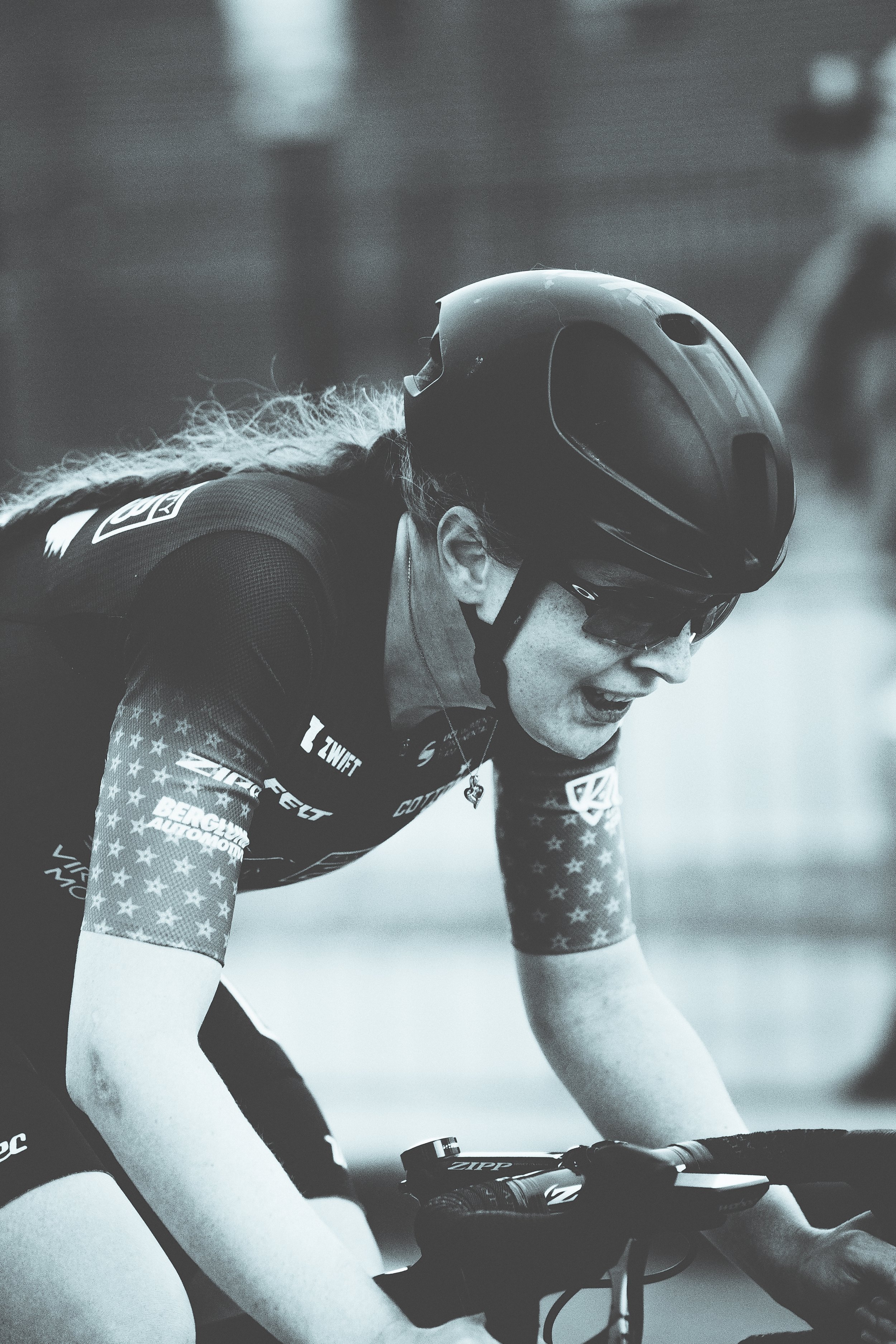 A woman cyclist wearing helmet, sunglasses, and a jersey with American flag design, intensely focused during a race.