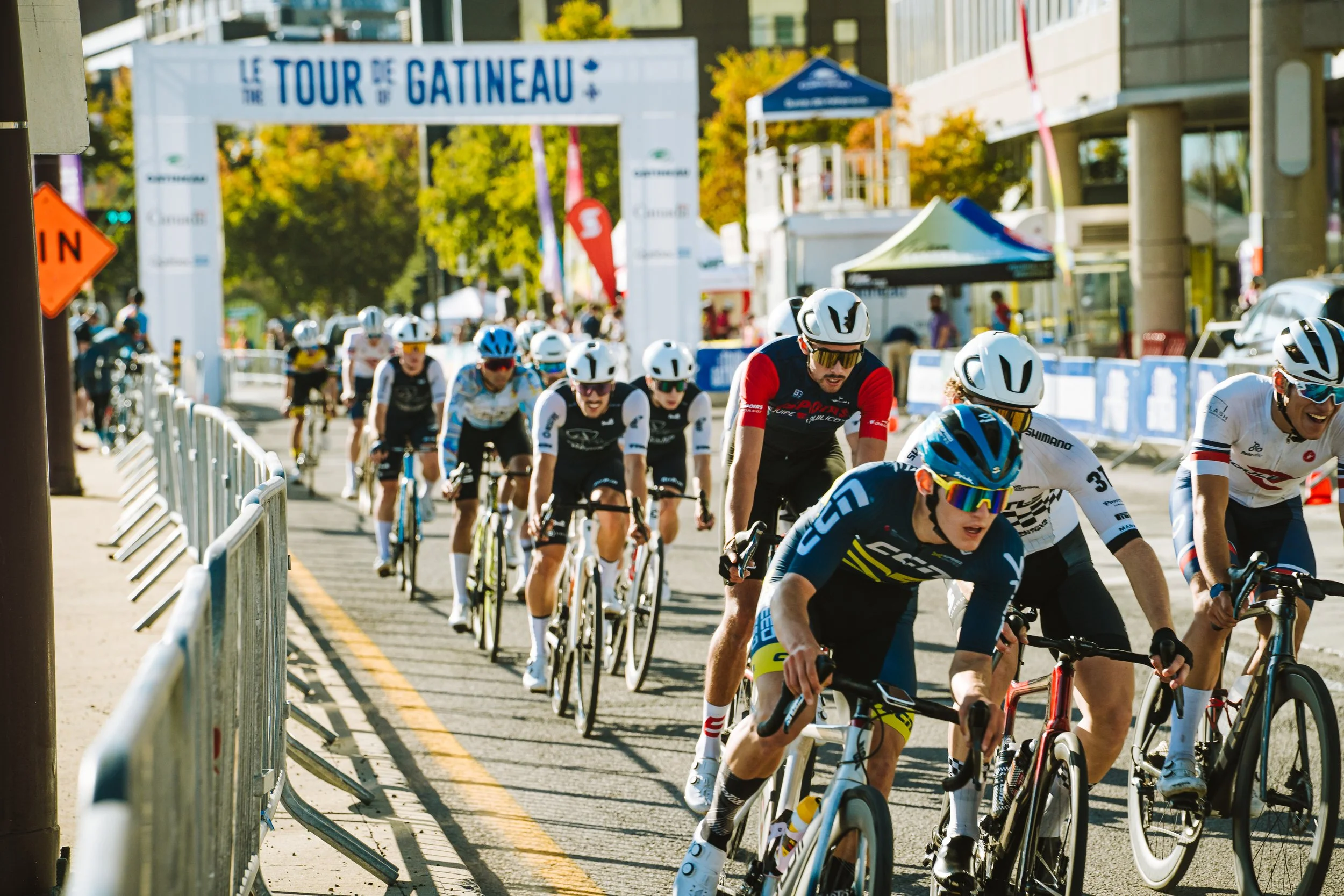 Cyclists in a competitive race riding on a city street lined with barriers, with trees and event banners in the background.