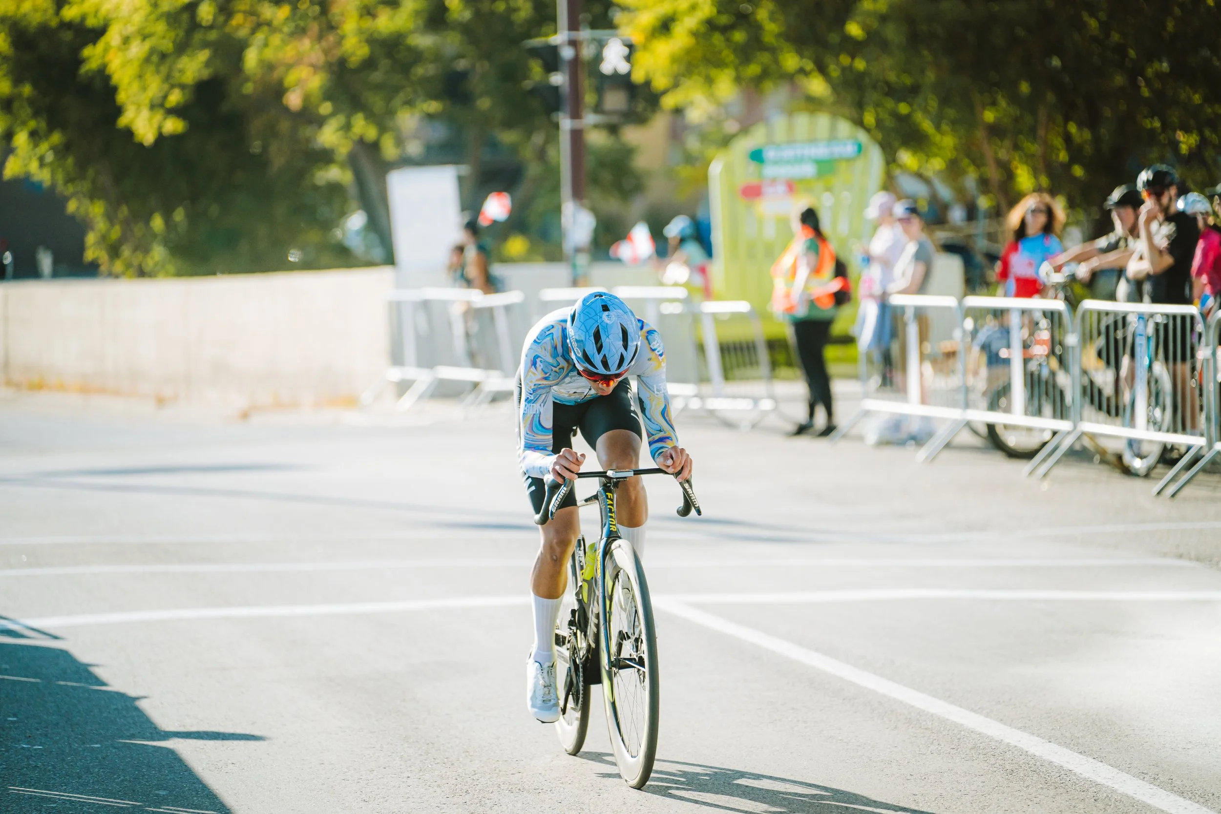 A cyclist in a blue helmet and colorful jersey riding a bicycle on a city street during a race, with spectators and race officials watching behind barriers.