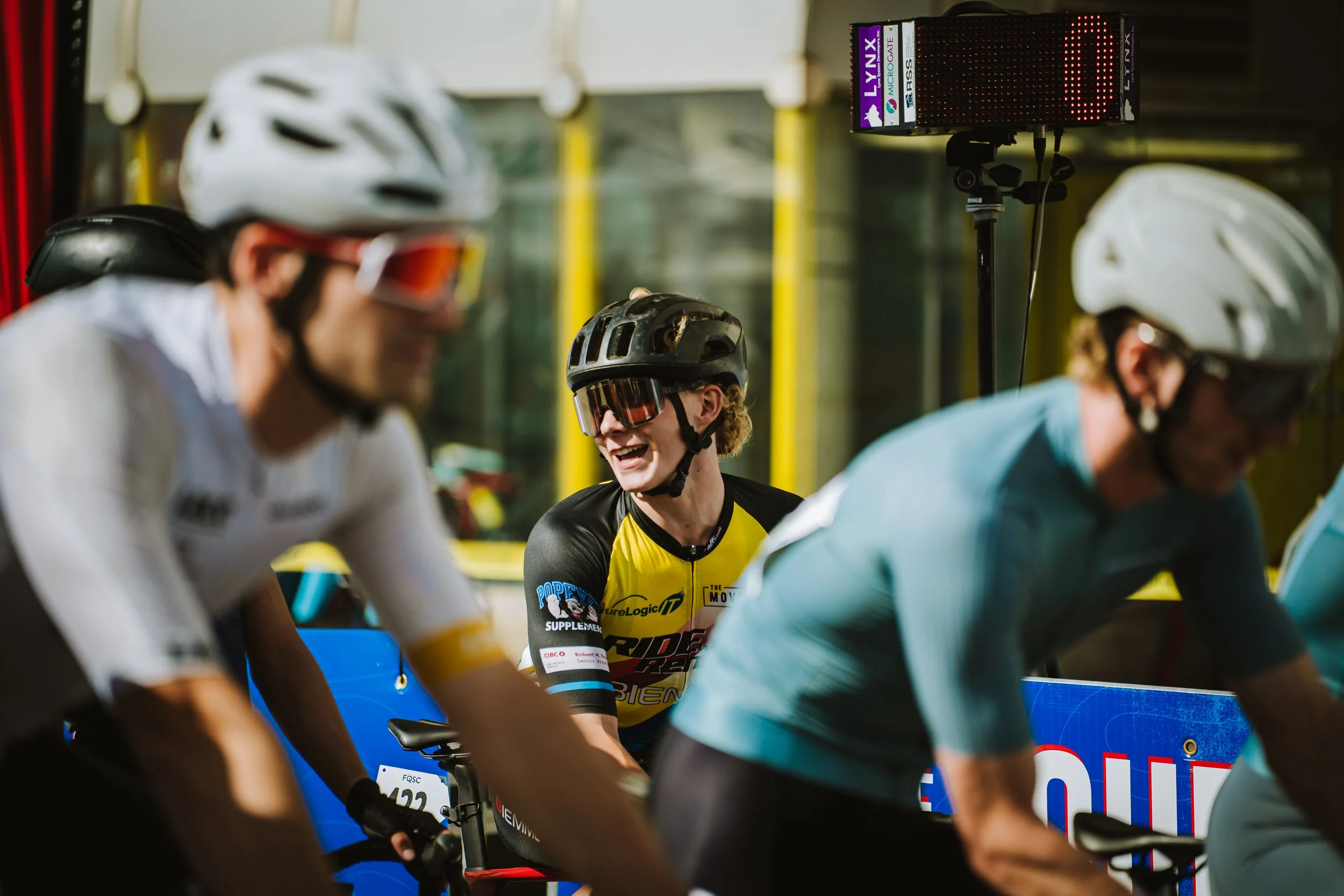 Three cyclists wearing helmets and sunglasses, with one smiling, on a track or in a velodrome, near a digital timer display. One cyclist has a yellow and black jersey, and the others are in white and blue jerseys.