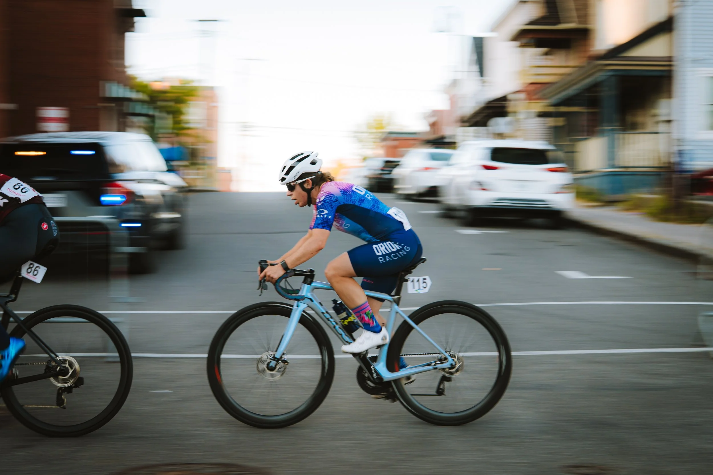 Cyclist wearing a blue and purple jersey and white helmet riding a blue bike on a city street during a race.