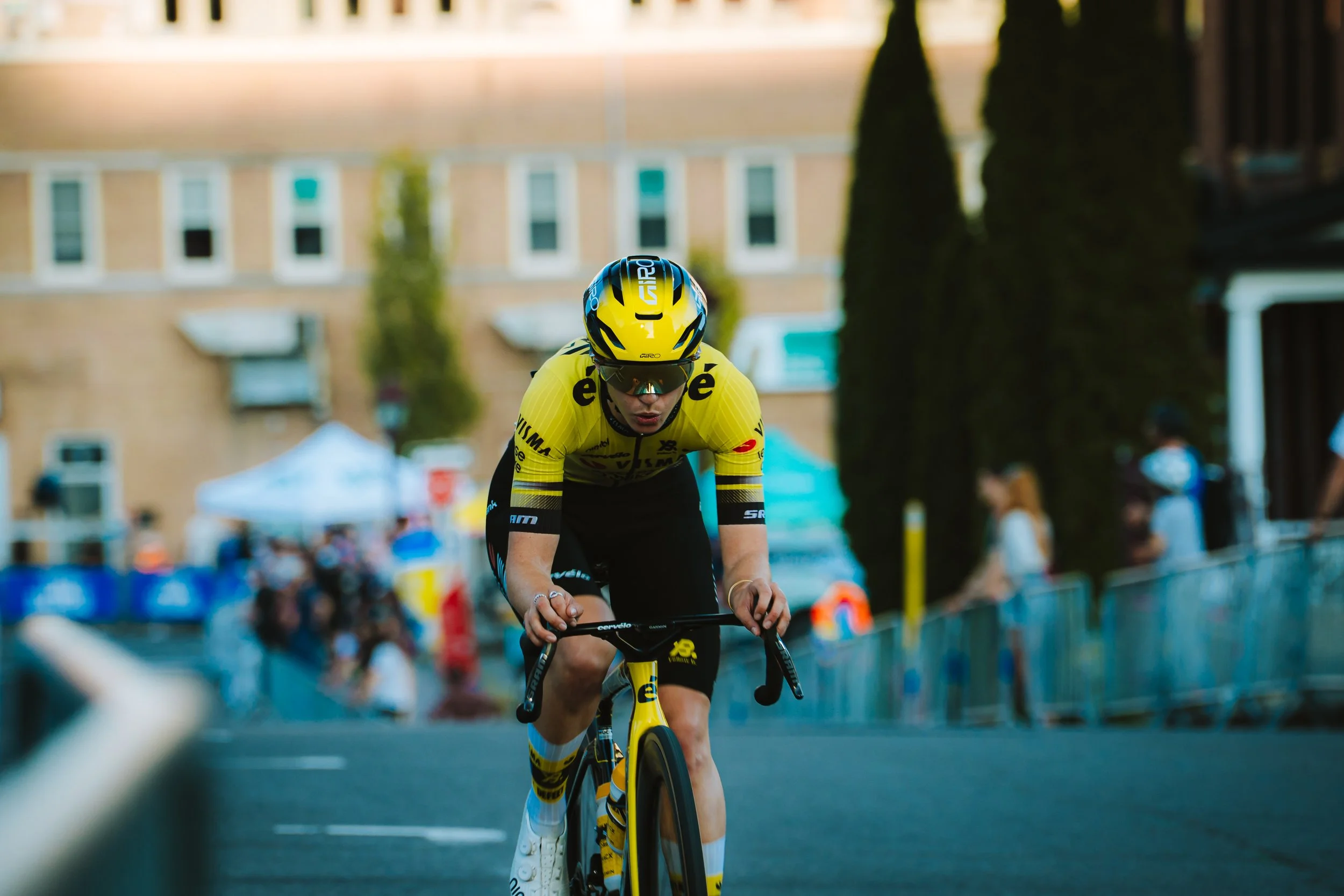 A cyclist in a yellow jersey and helmet riding in a race, leaning forward on a race bike, with a crowd and tents in the background.