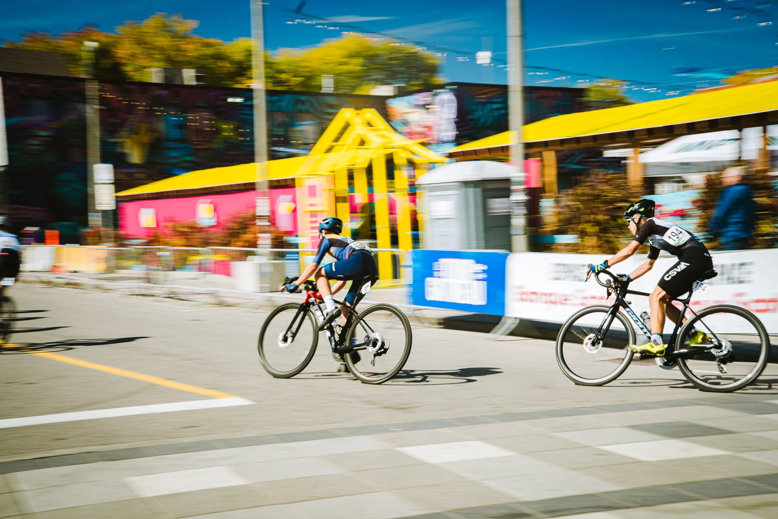 Two cyclists in a race, riding on a city street with colorful buildings and banners in the background, motion blur indicating speed.