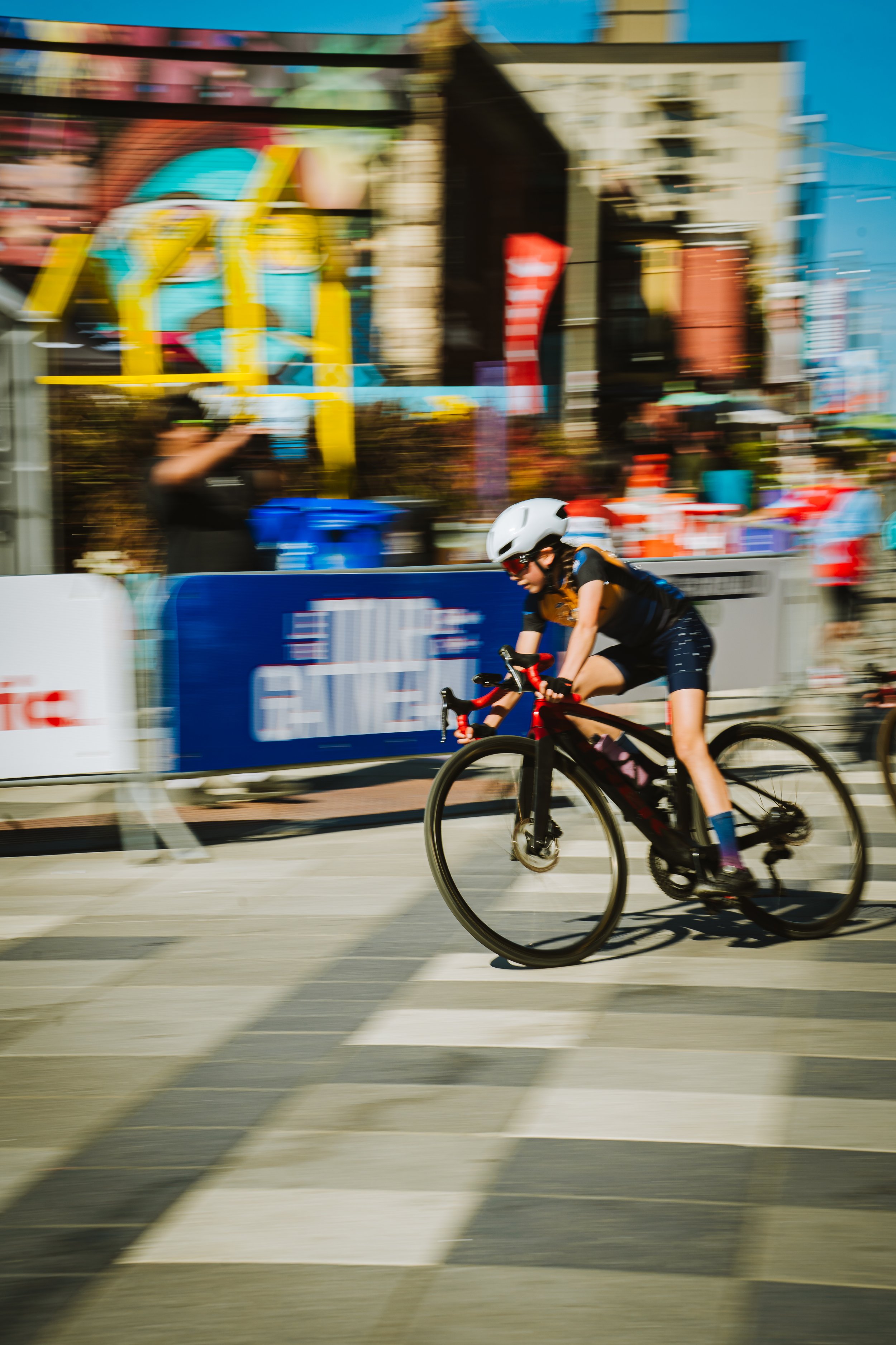 A female cyclist wearing a white helmet and black outfit racing on a city street, blurred background indicating motion.
