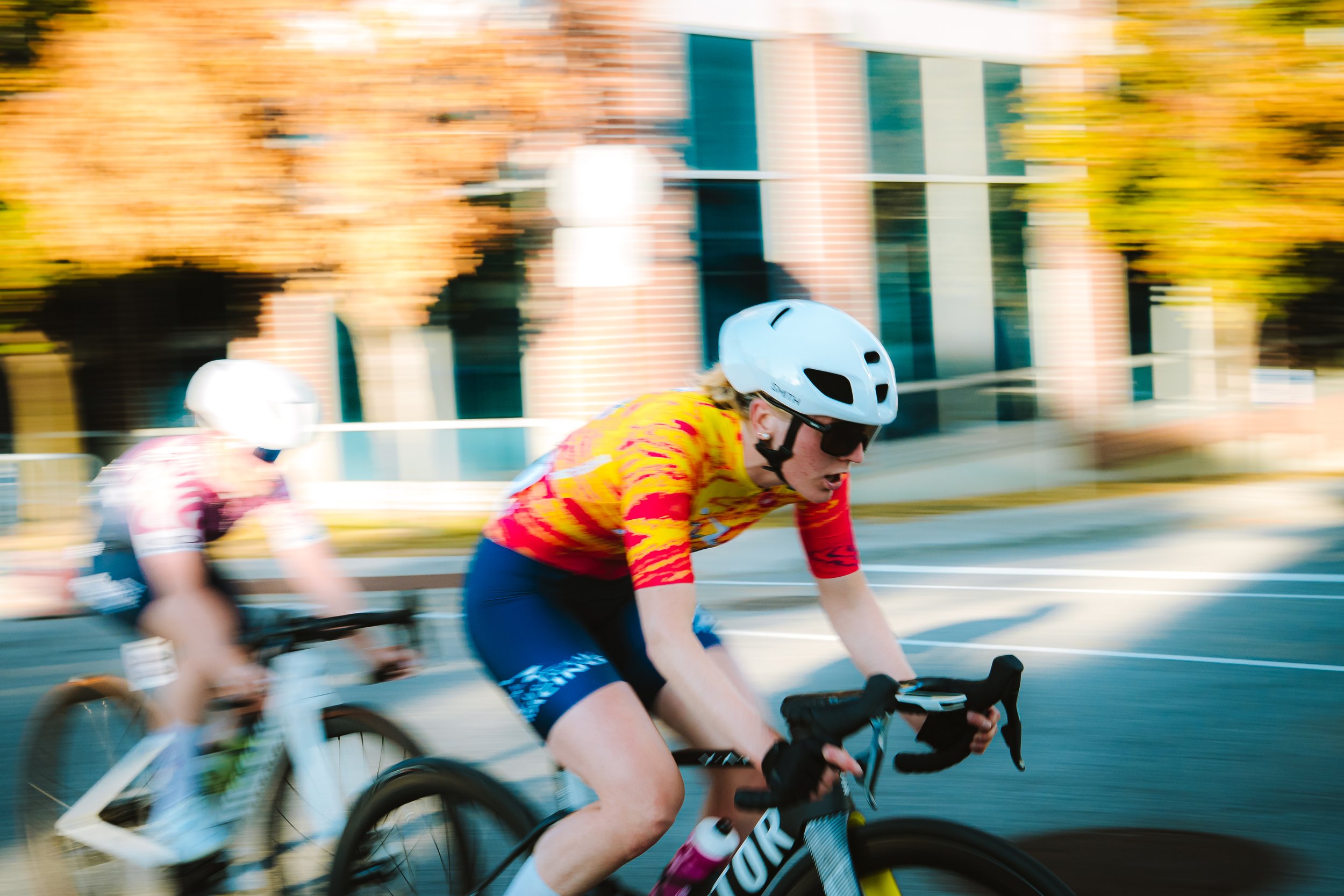 Cyclist in yellow and red jersey riding a black bike with white helmet and sunglasses, blurred background of trees and a building