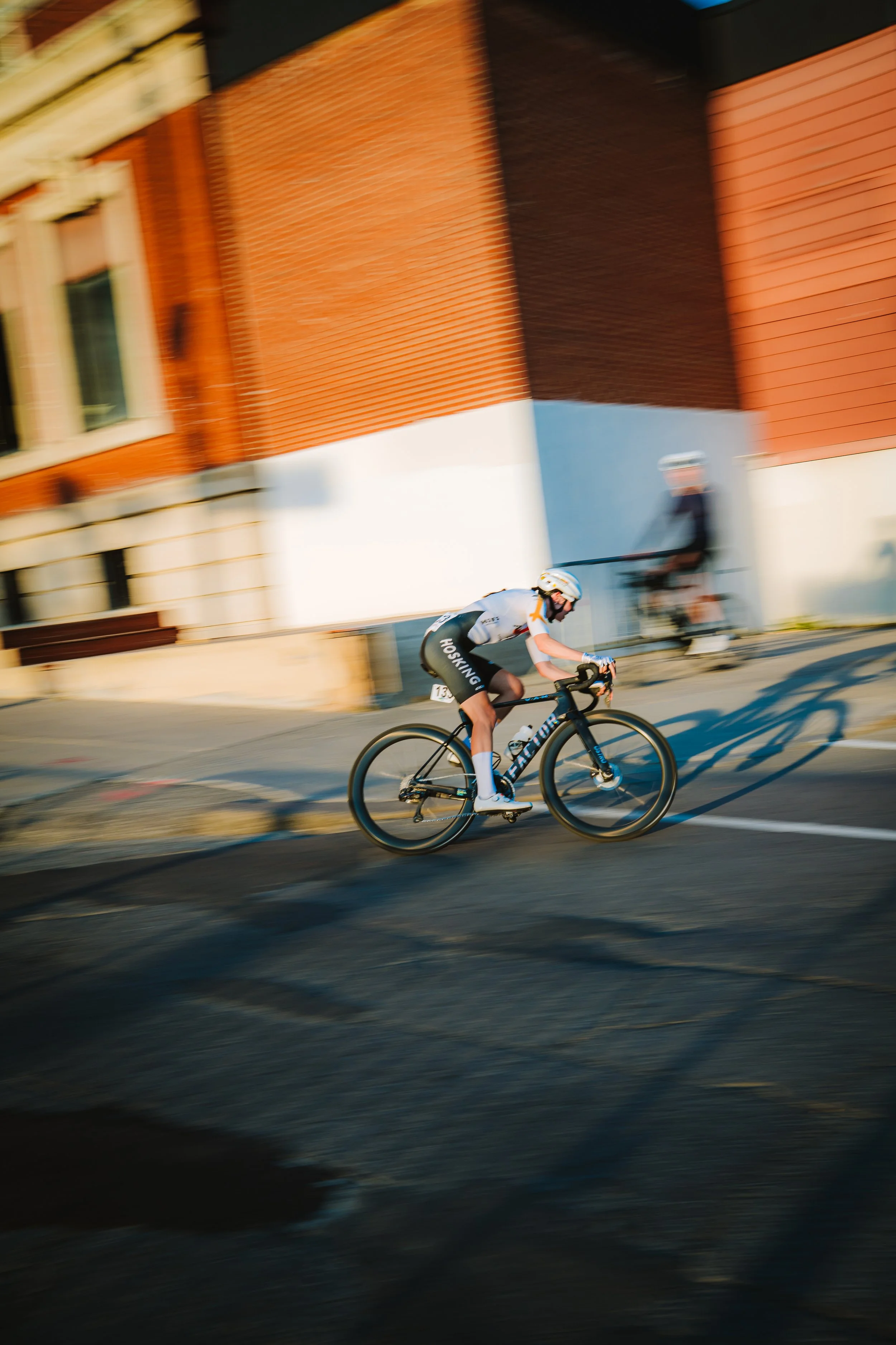 A cyclist racing on a city street with a blurred background of buildings and another person in the background.